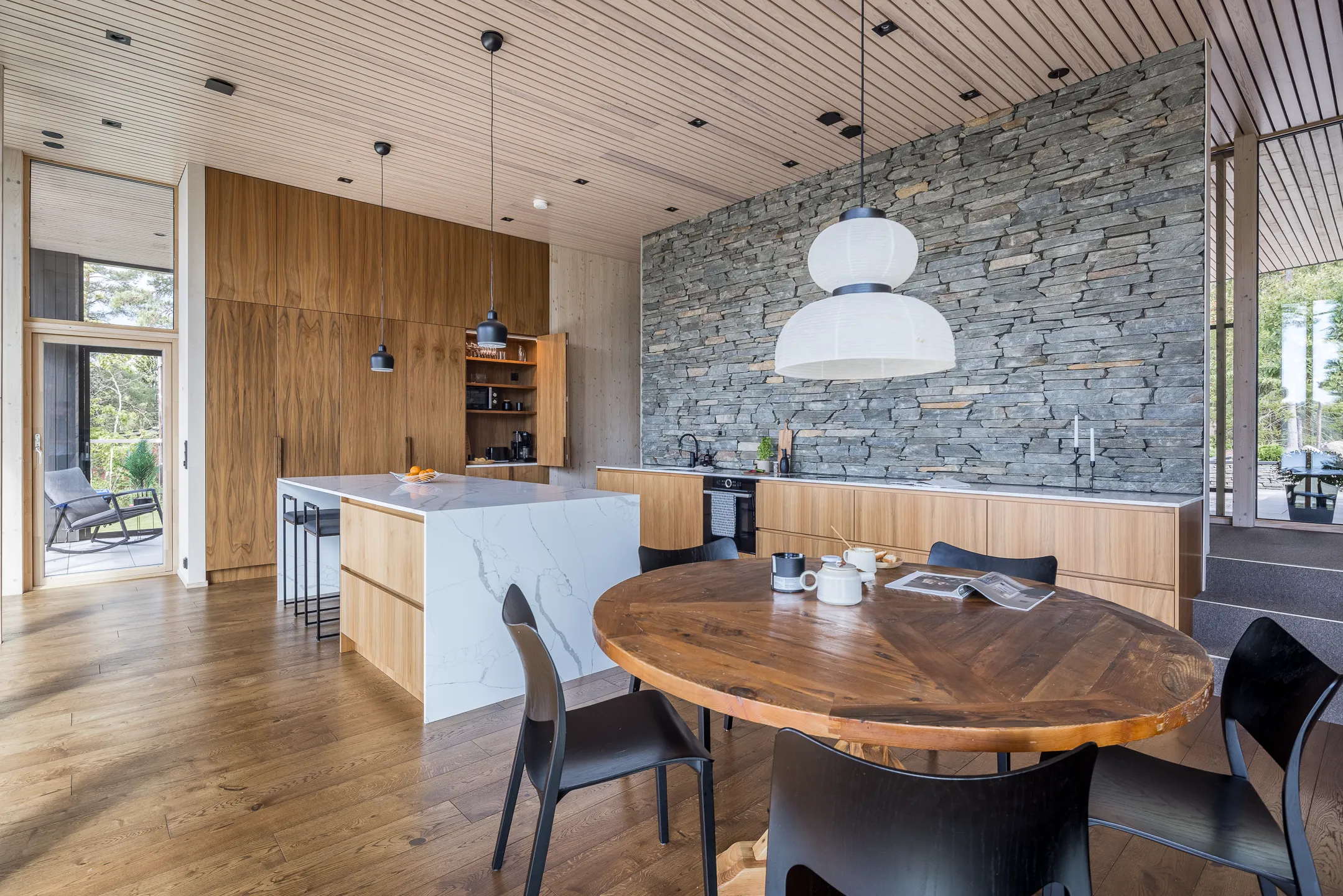 Moderni keittiö, jossa on puulattia, kiviseinä, puukaapit, marmorinen keittiösaareke ja pyöreä puinen ruokapöytä mustilla tuoleilla.