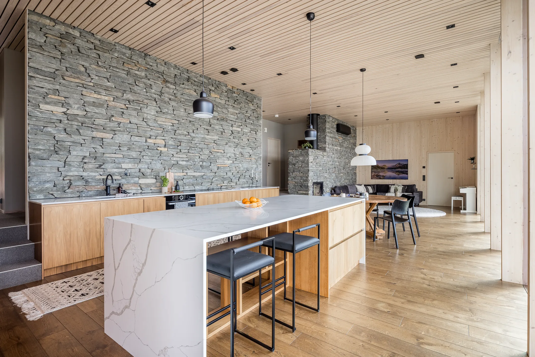 Moderni sisustus kiviseinällä, puukatto ja -lattia, keittiösaareke ja mustat jakkarat valoisassa tilassa.