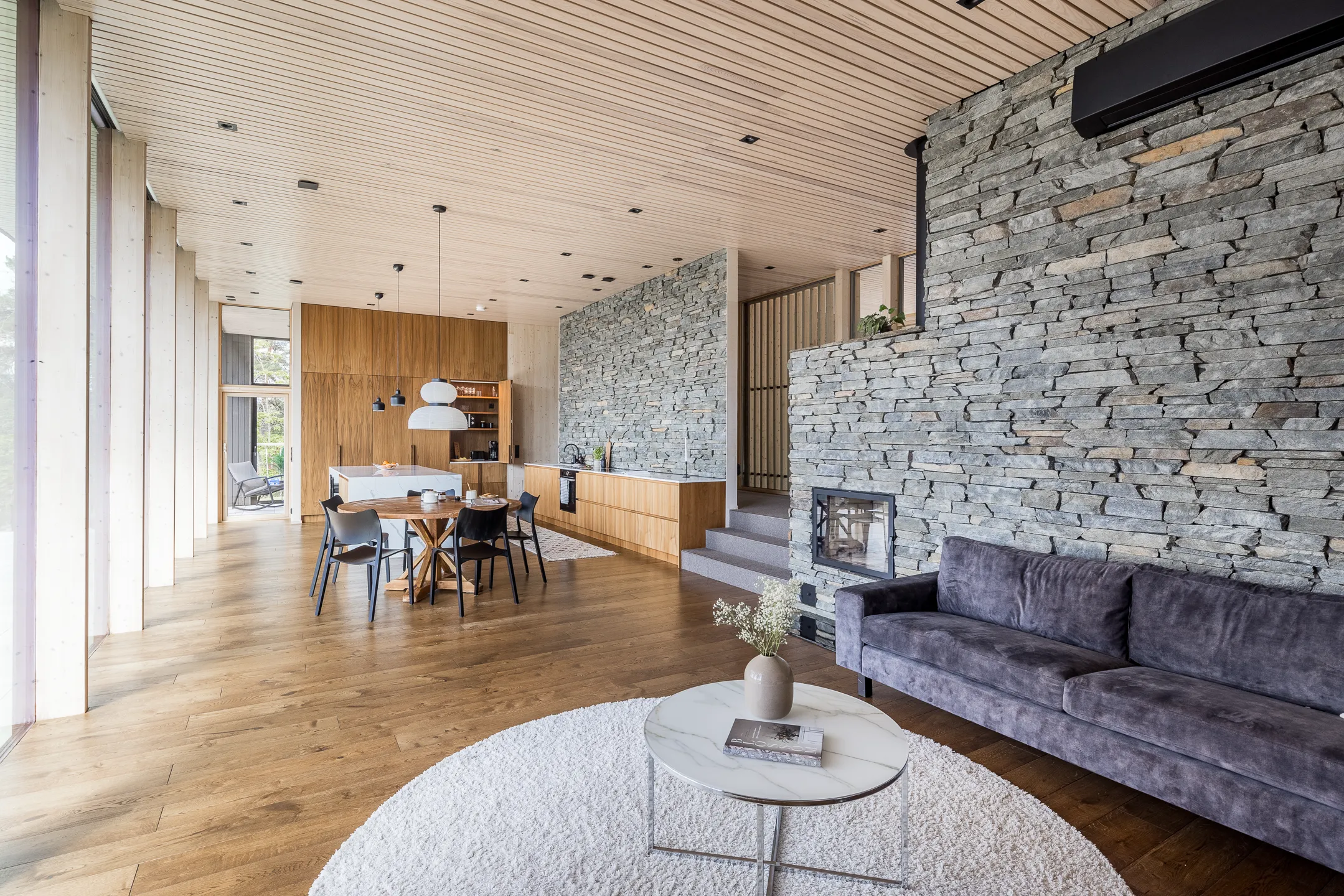 Valoisa ja moderni sisustus: kiviseinä, harmaa sohva, pyöreä matto, puukatto ja -lattia.