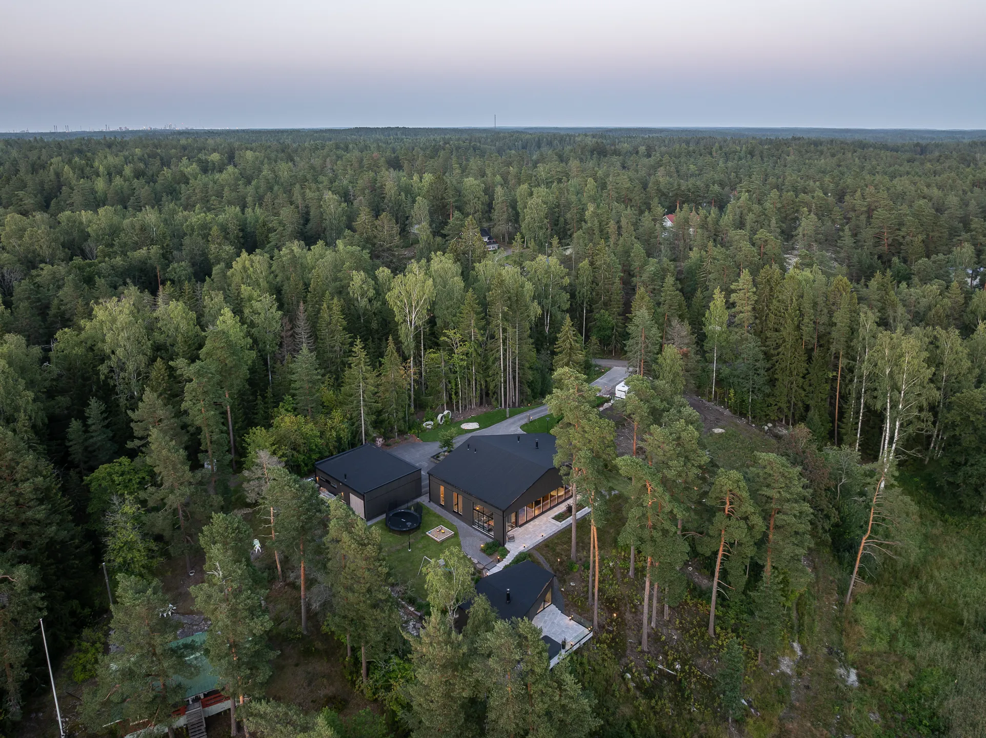 Ilmakuva modernista mustasta talosta metsän keskellä, jossa on vihreitä puita ja harmaa taivas.