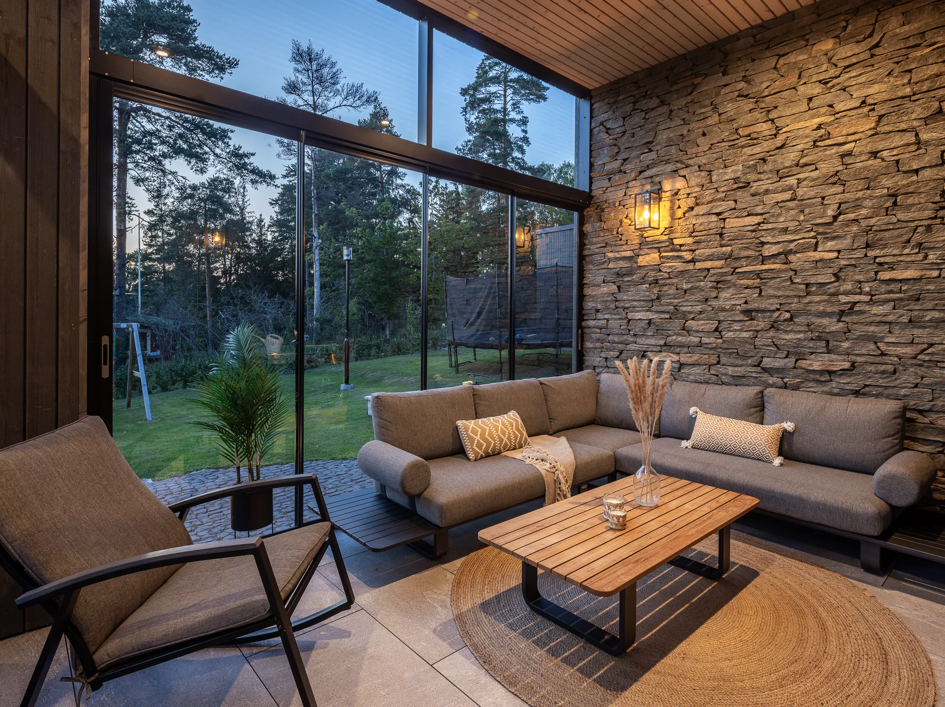 Valoisa olohuone kiviseinällä, puukatto, suuri ikkuna, harmaa sohva, puinen pöytä ja pyöreä matto.