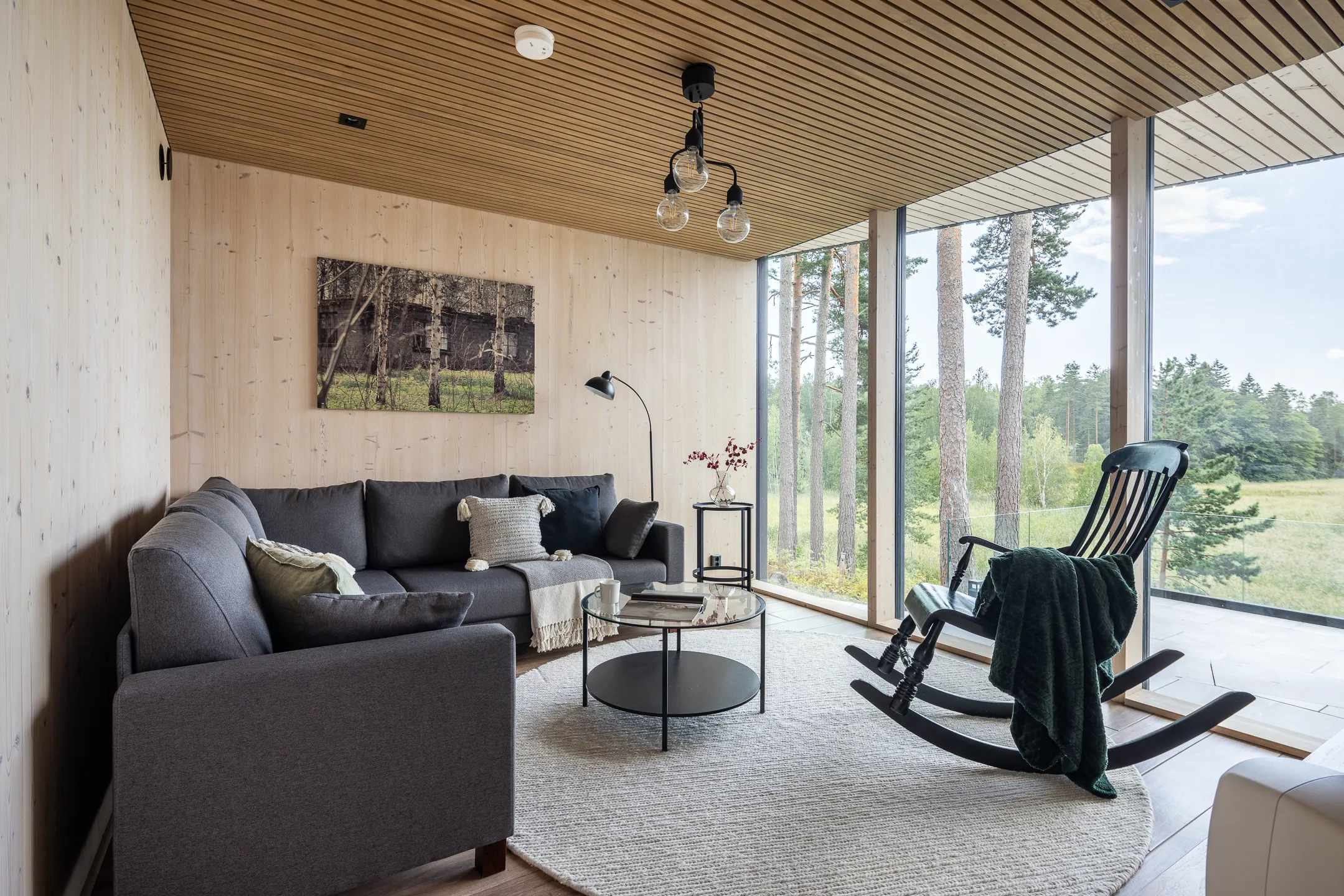 Valoisassa olohuoneessa on harmaa kulmasohva, pyöreä matto, musta keinutuoli ja suuret ikkunat, joista näkyy metsämaisema. Puupanelointi seinissä ja katossa luo lämpimän tunnelman.
