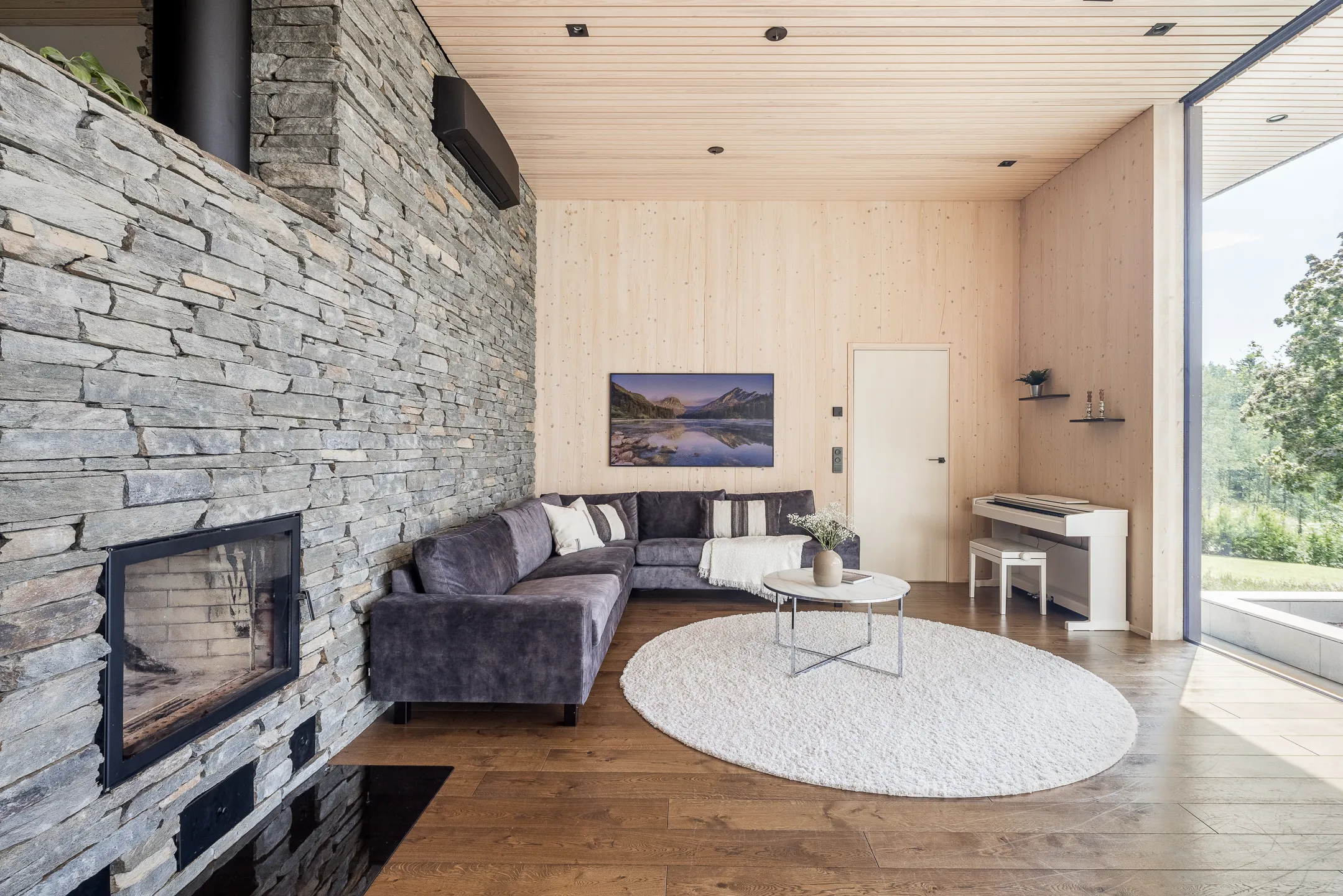 Olohuone kiviseinällä, harmaalla sohvalla, valkoisella matolla ja puupaneeliseinillä, jossa on piano ja taulu.