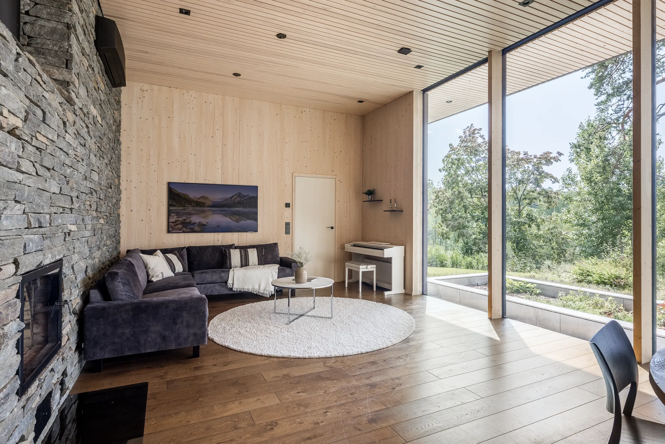 Olohuoneessa on kiviseinä, tumma sohva, valkoinen pyöreä matto, puinen katto ja seinät sekä suuret ikkunat, joista näkyy vehreä maisema.