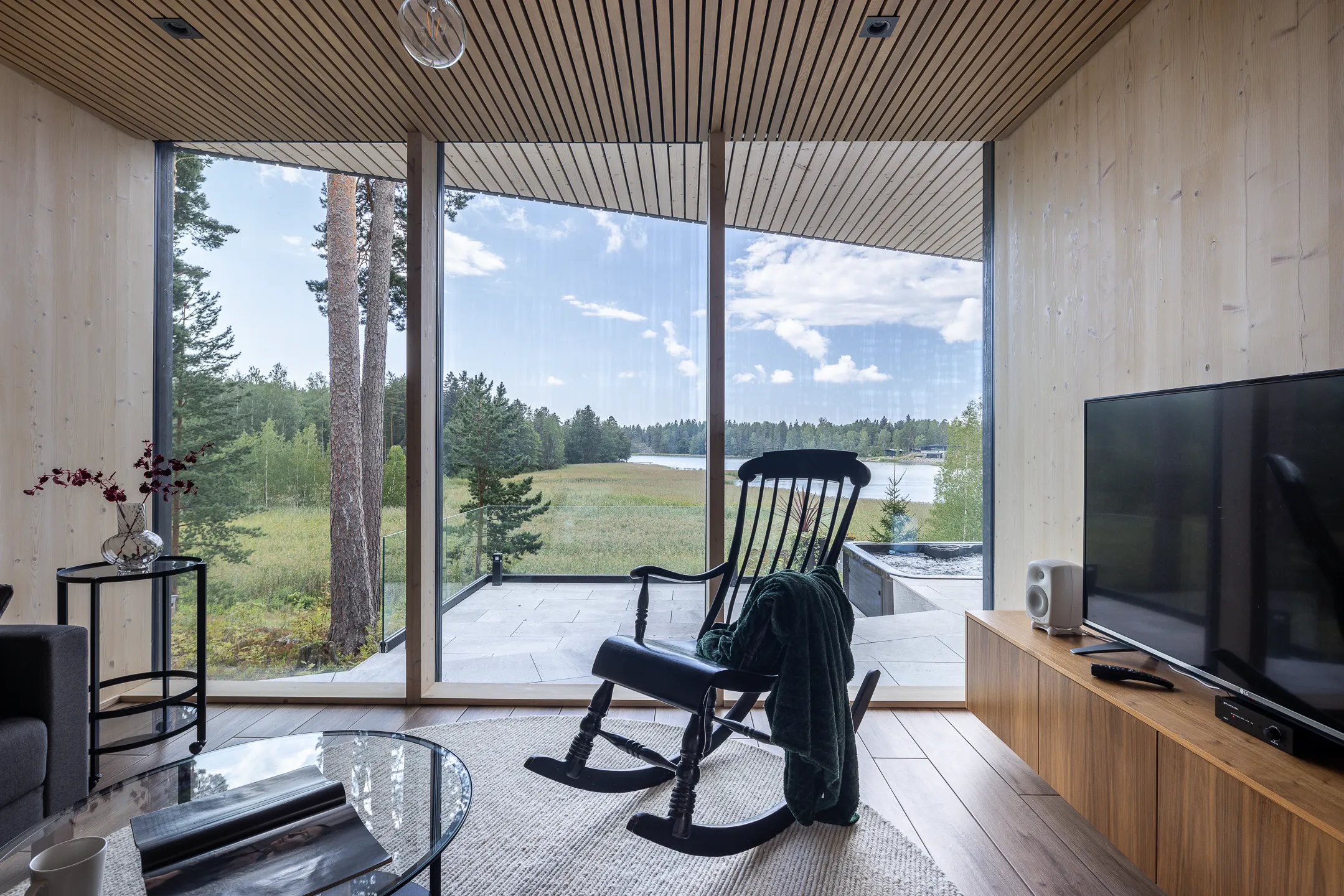 Valoisassa olohuoneessa on suuri ikkuna, jonka edessä on musta keinutuoli ja vihreä viltti. Huoneessa on myös sohva, sohvapöytä, sivupöytä ja TV-taso.