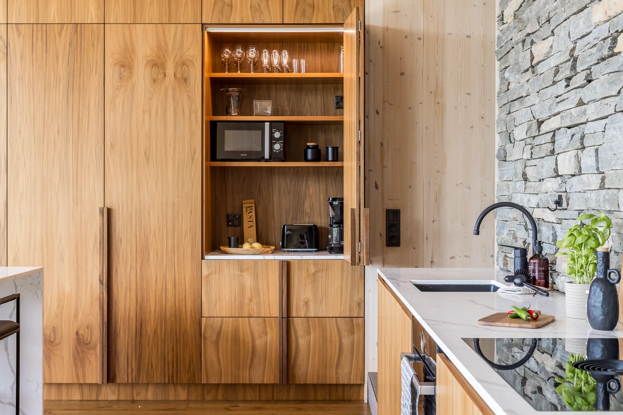 Puinen keittiö, jossa on avoin kaappi, jossa on viinilaseja, mikroaaltouuni ja kahvinkeitin, sekä kiviseinä ja valkoinen työtaso.