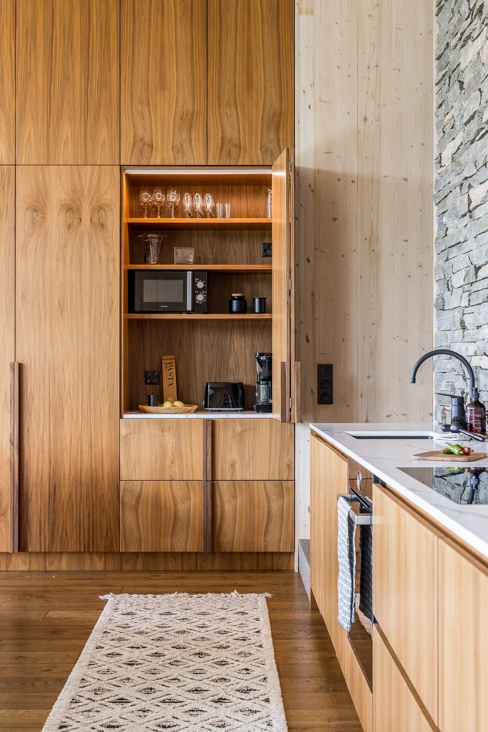 Puinen keittiö, jossa on avoin kaappi, jossa on lasit, mikroaaltouuni ja kahvinkeitin, sekä valkoinen matto lattialla.