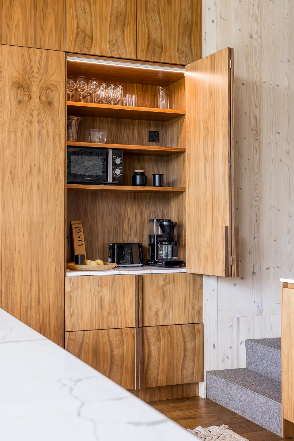 Puinen keittiökaappi avoimella ovella, sisältää lasitavaraa, mikroaaltouunin, kahvinkeittimen ja muita keittiötarvikkeita, valaistus ylhäällä.