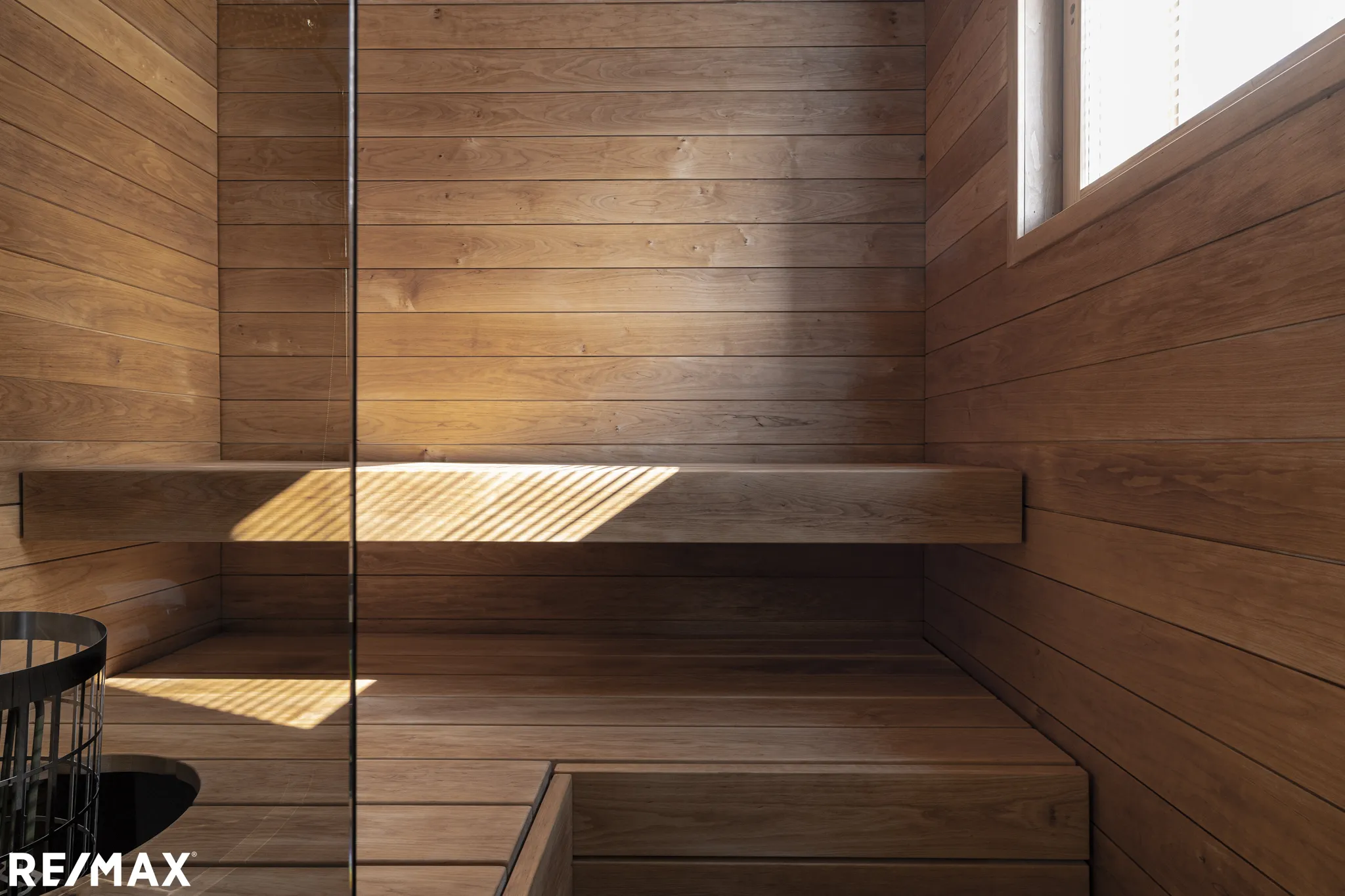 Puinen sauna, jossa on penkit, ikkuna ja RE/MAX-logo alhaalla vasemmalla, valaistu auringonvalolla.