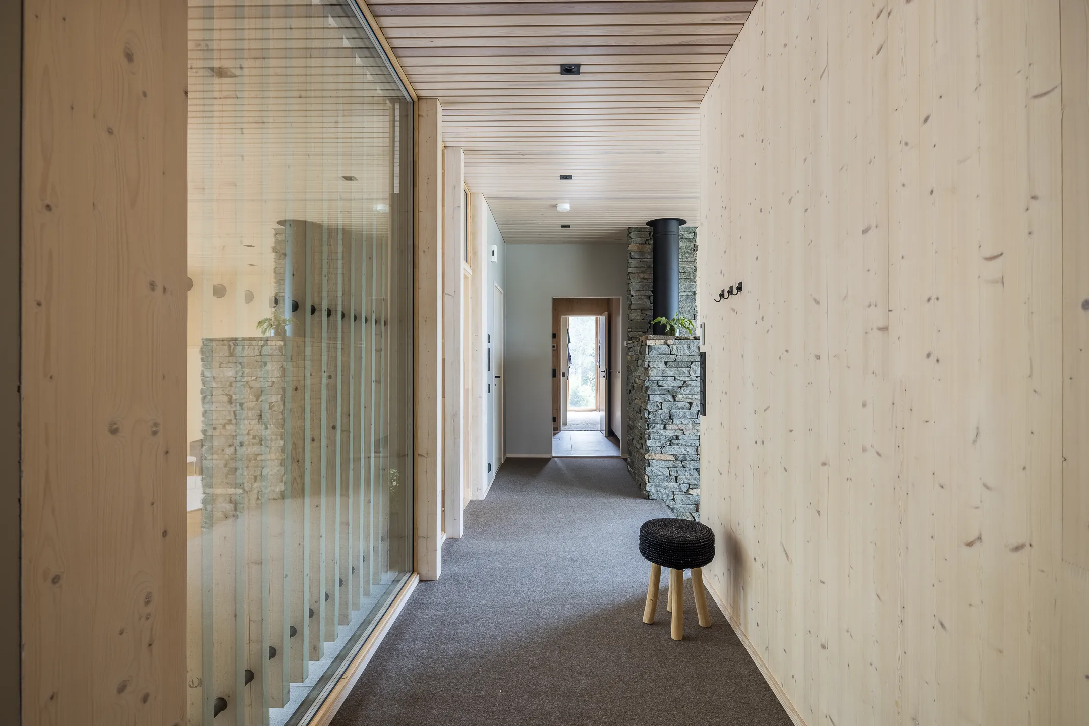 Vaalean puun sisustus, harmaa matto, musta jakkara ja kiviseinäinen takka valoisassa käytävässä.