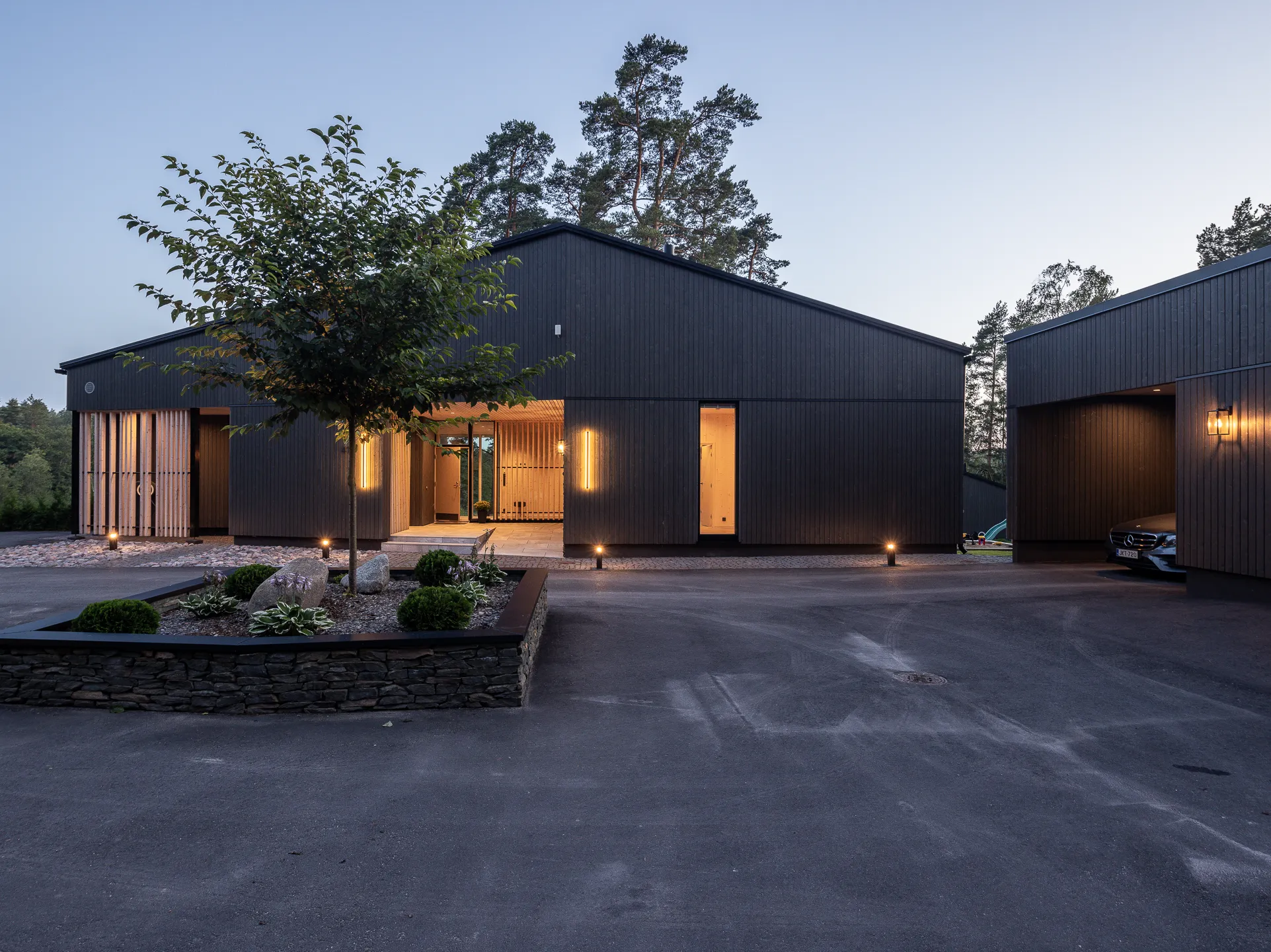 Moderni, tumma puutalo hämärässä, jossa on valaistu etupiha, kivikkopuutarha ja autokatos, luoden tyylikkään ja kutsuvan tunnelman.