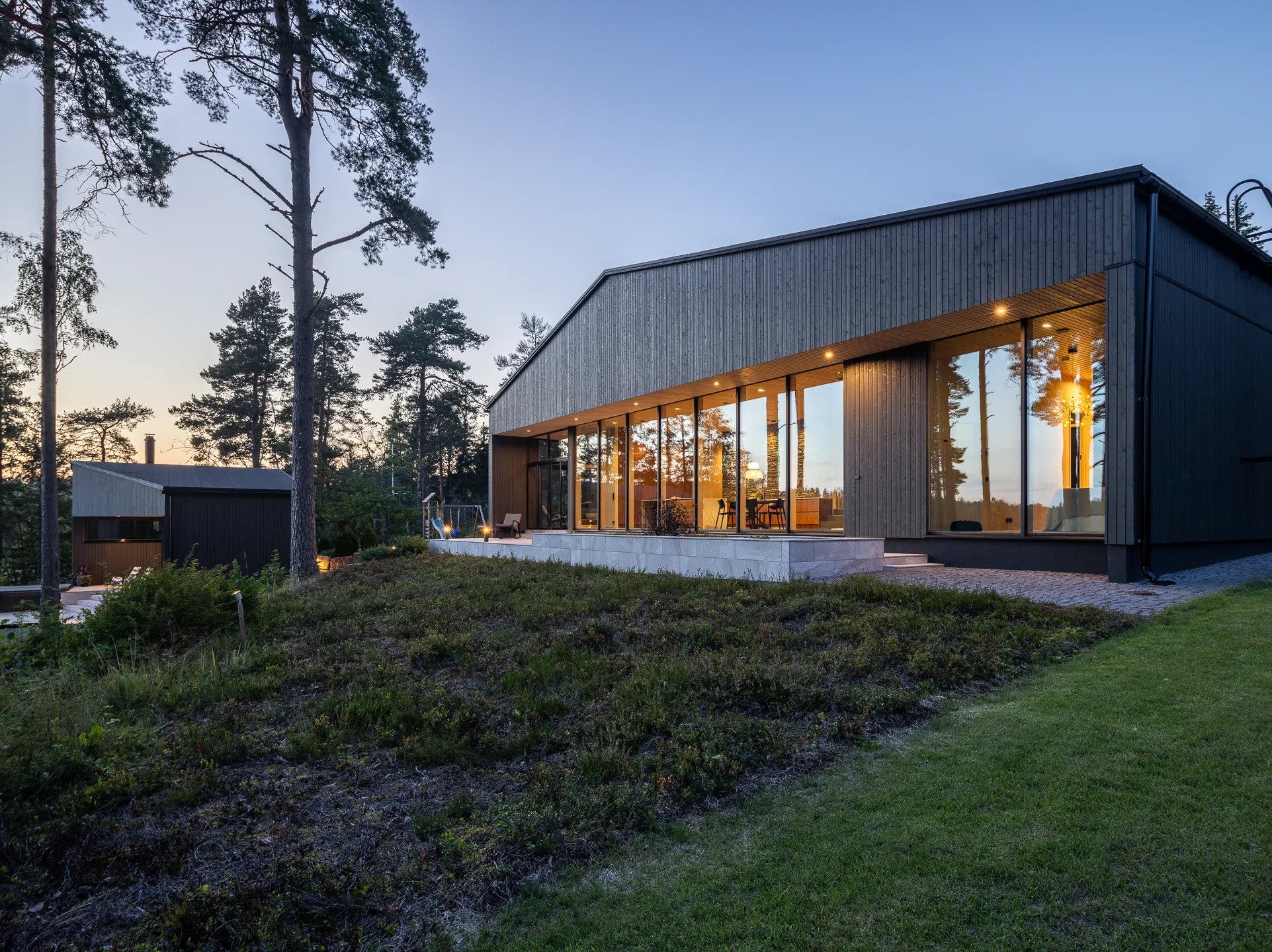 Moderni puutalo iltavalaistuksessa, suuret ikkunat heijastavat sisätiloja ja ympäröivää luontoa, tumma puuverhous ja vihreä ympäristö.