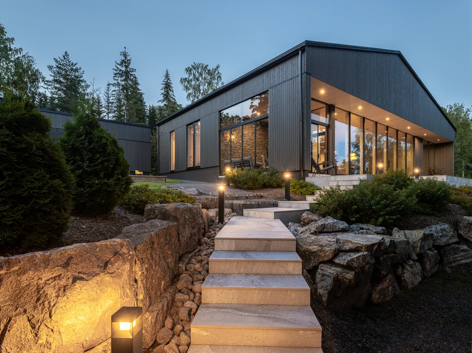 Moderni, tumma puutalo kivipolulla ja valaistuksella iltahämärässä.