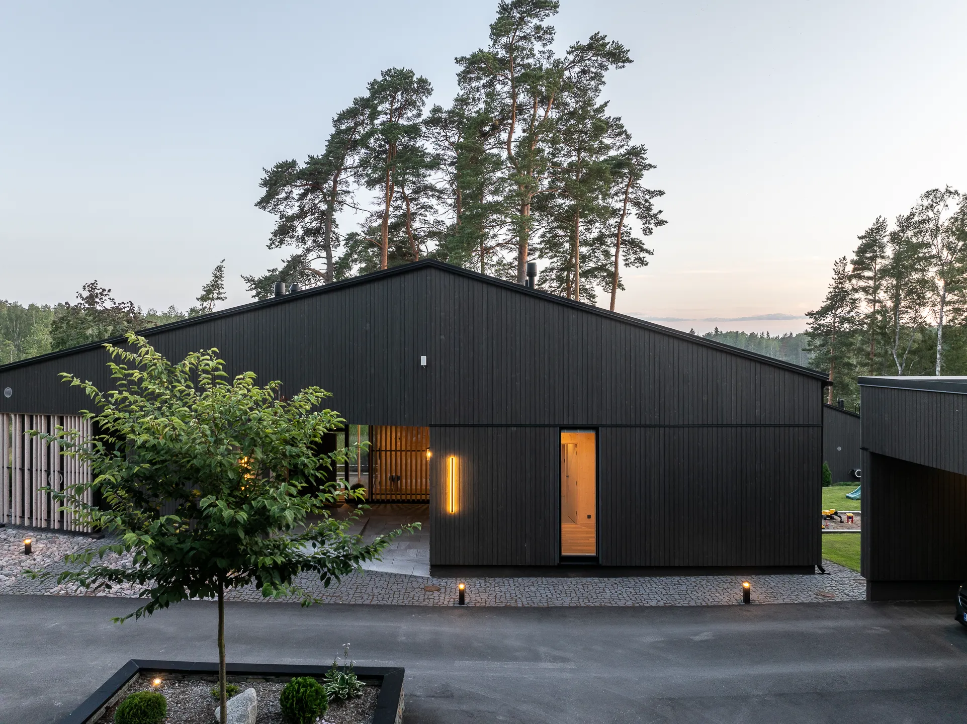 Moderni, tumma puutalo, jossa on valaistu sisäänkäynti ja pihalla nuori puu. Taustalla näkyy korkeita mäntyjä ja hämärtyvä taivas.