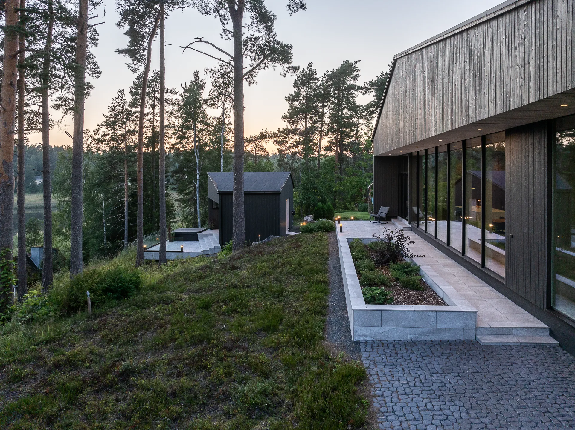 Moderni, tumma puutalo metsän reunalla, jossa suuret ikkunat ja kivetty piha. Pihalla on istutusalueita ja polkuja, ja taustalla näkyy metsää ja järvi. Valo on pehmeää ja iltaista.