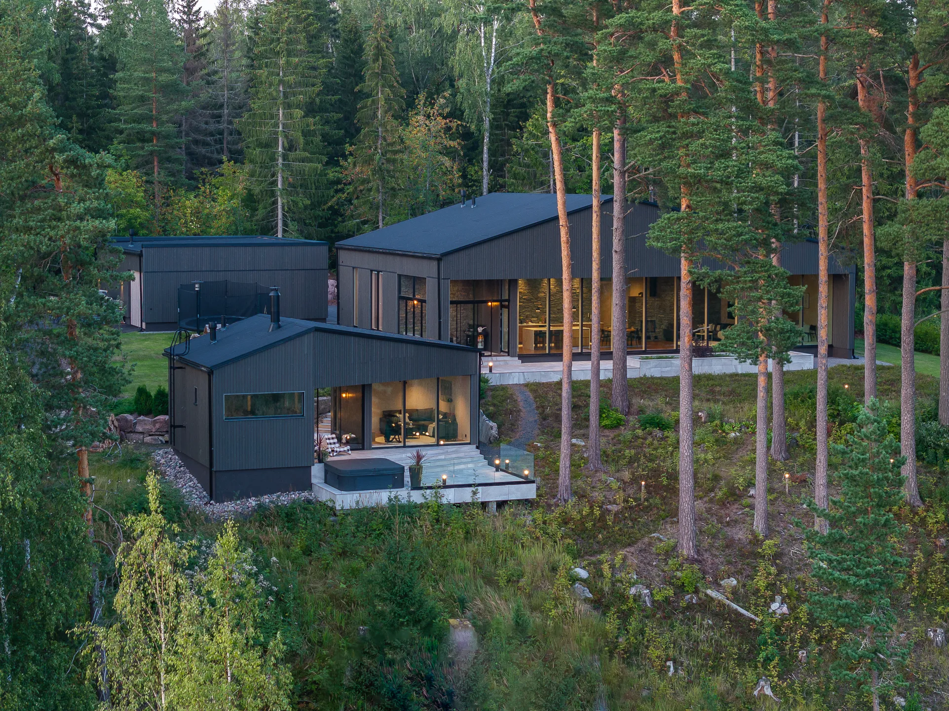 Moderni, tumma puutalo metsäisessä ympäristössä, jossa on suuret ikkunat ja ulkoterassi poreammeella.