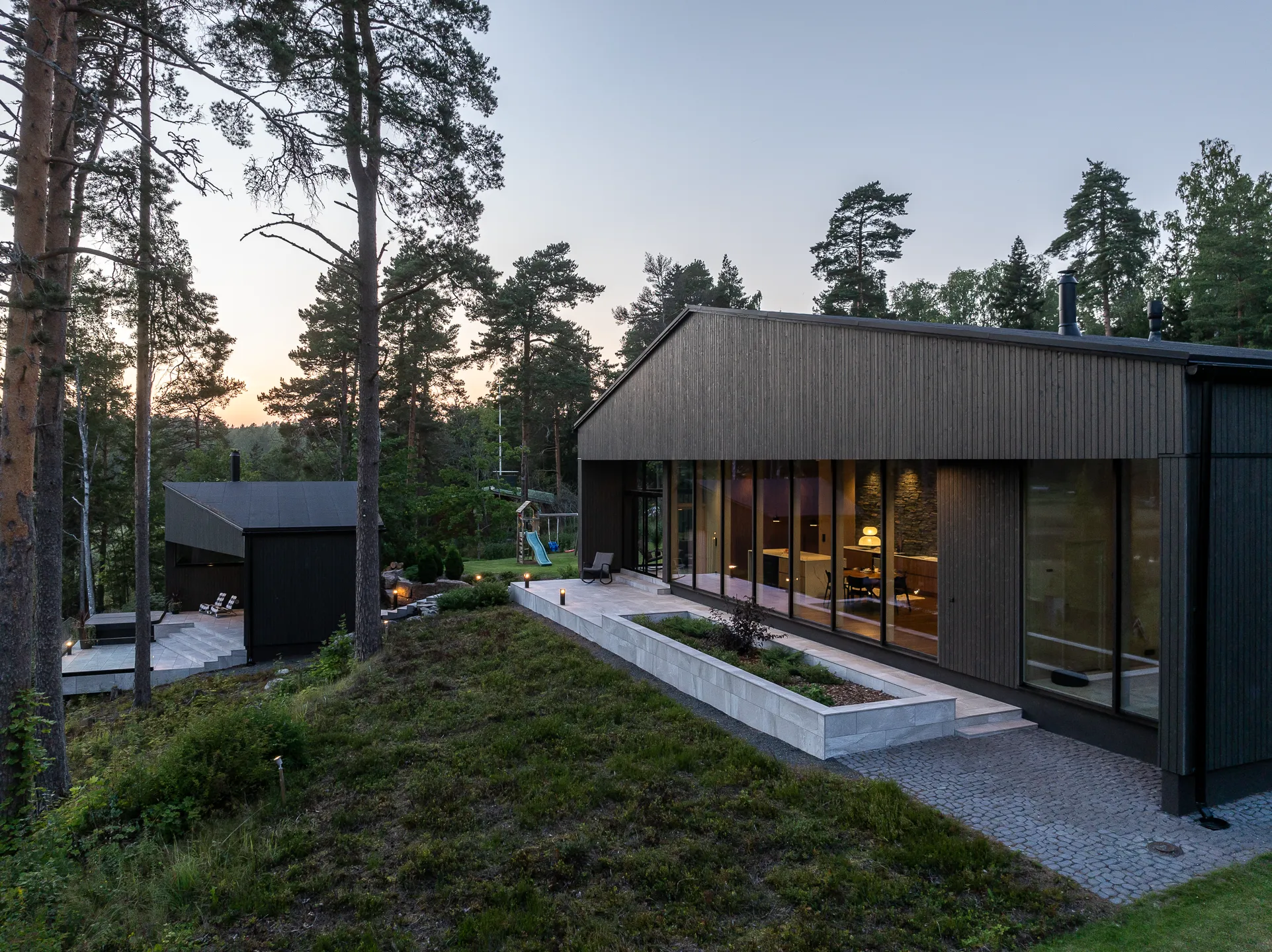 Moderni, tumma puutalo metsäisessä maisemassa iltavalaistuksessa, jossa suuret ikkunat heijastavat sisätilojen valoa ja ulkona on kivetty polku ja istutusalue.