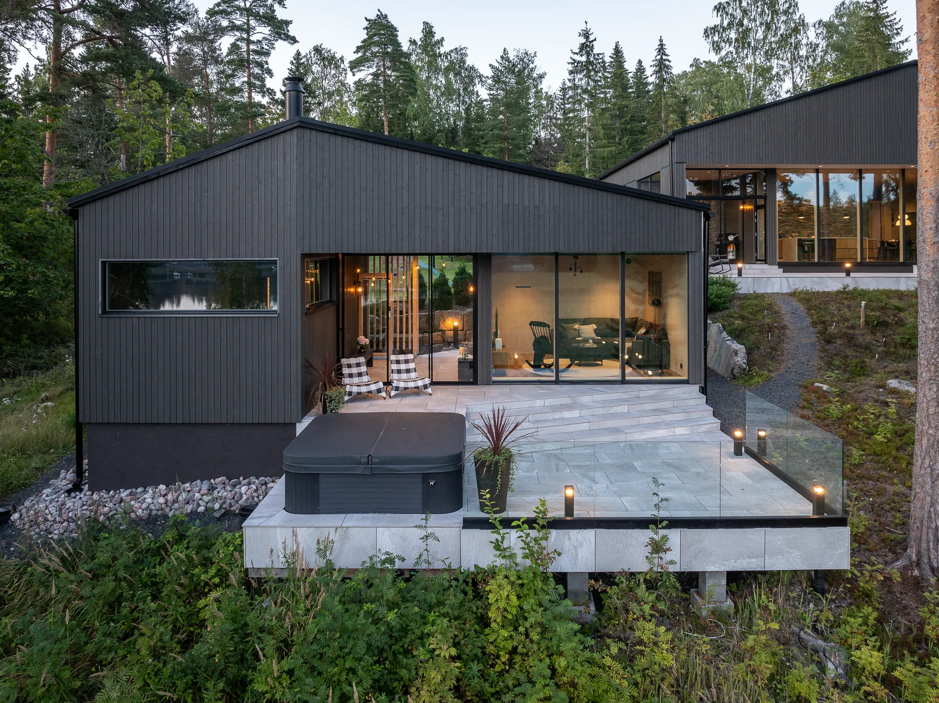 Moderni, tumma puutalo, jossa suuret ikkunat, terassi ja poreallas, ympäröitynä metsällä ja vihreällä kasvillisuudella.