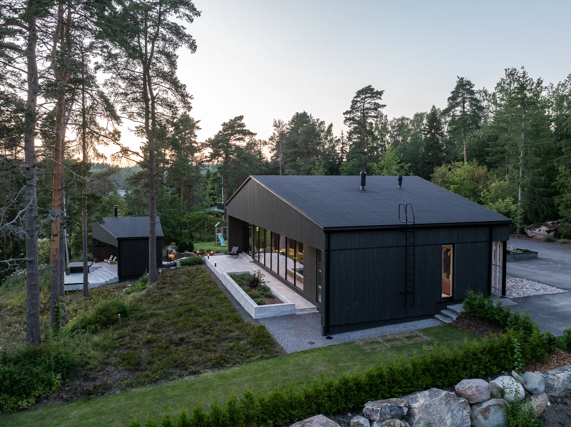 Moderni, tumma puutalo metsäisessä ympäristössä, jossa on suuret ikkunat, tasainen katto ja erillinen pienempi rakennus. Maisemassa on kiviä, nurmikkoa ja puita.