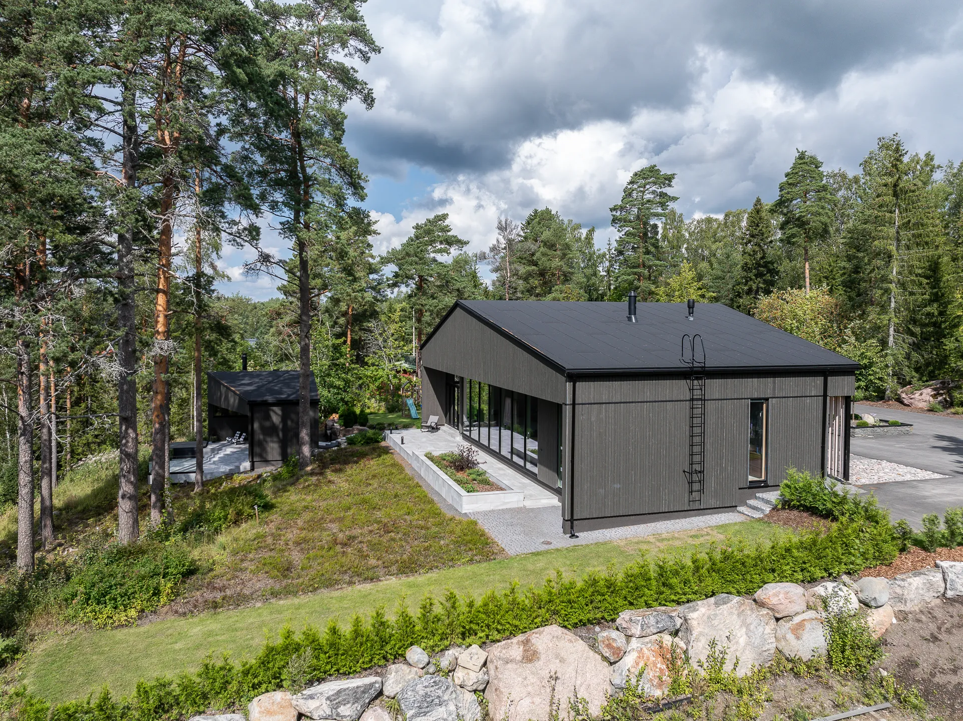 Moderni tumma puutalo metsäisessä maisemassa, jossa on harmaa katto, suuret ikkunat ja kivimuuri etualalla, valoisa sää.