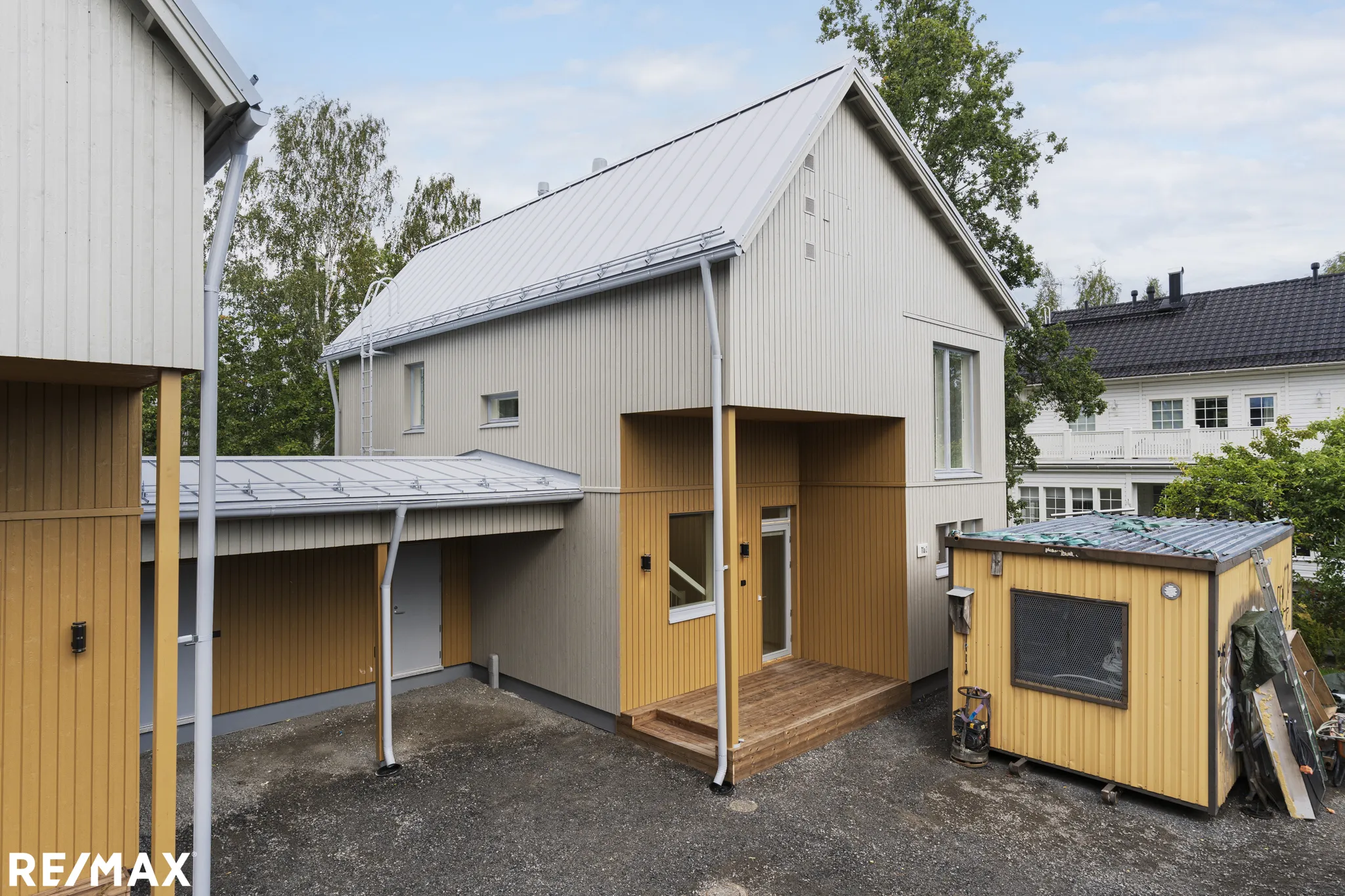Moderni, kaksikerroksinen puutalo, jossa on harmaa ulkovuoraus ja keltainen korostus, sekä pieni piharakennus ja RE/MAX-logo alhaalla.