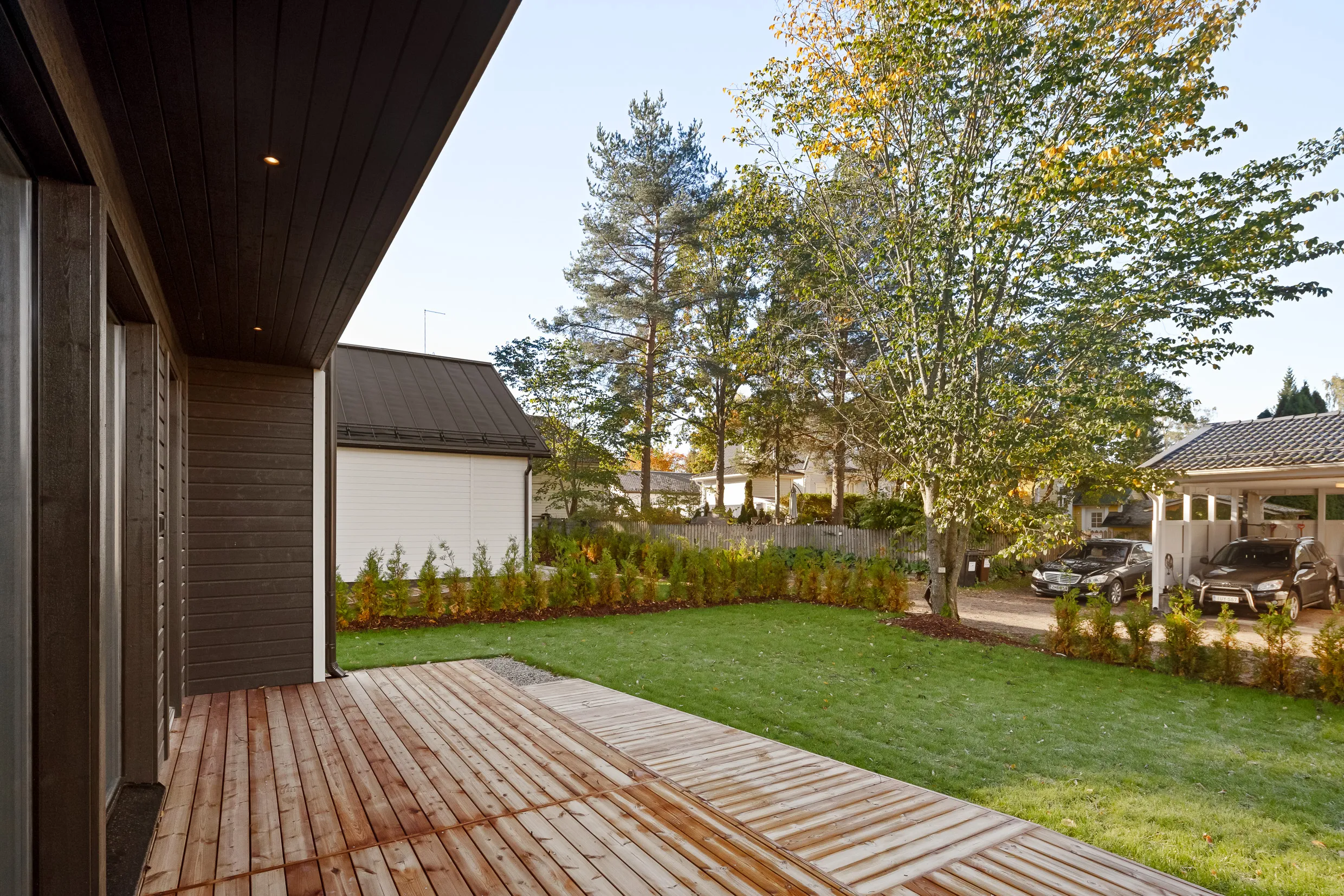 Ulkoterassi, jossa on puinen kansi, vihreä nurmikko ja puita. Taustalla näkyy talo ja autokatos, jossa on kaksi autoa. Valoisa ja aurinkoinen sää.