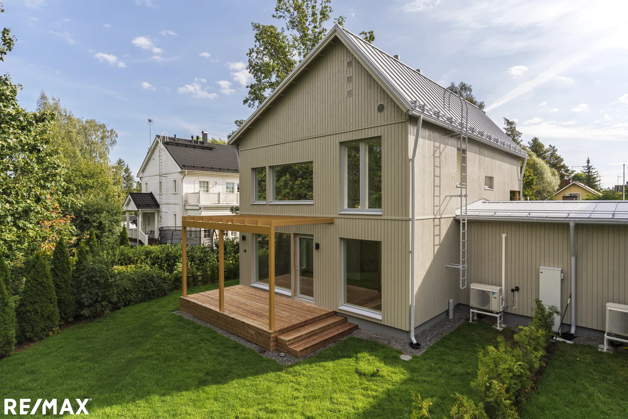 Valokuva ulkoa modernista, kaksikerroksisesta puutalosta, jossa on puinen terassi ja metallikatto, aurinkoisena päivänä.