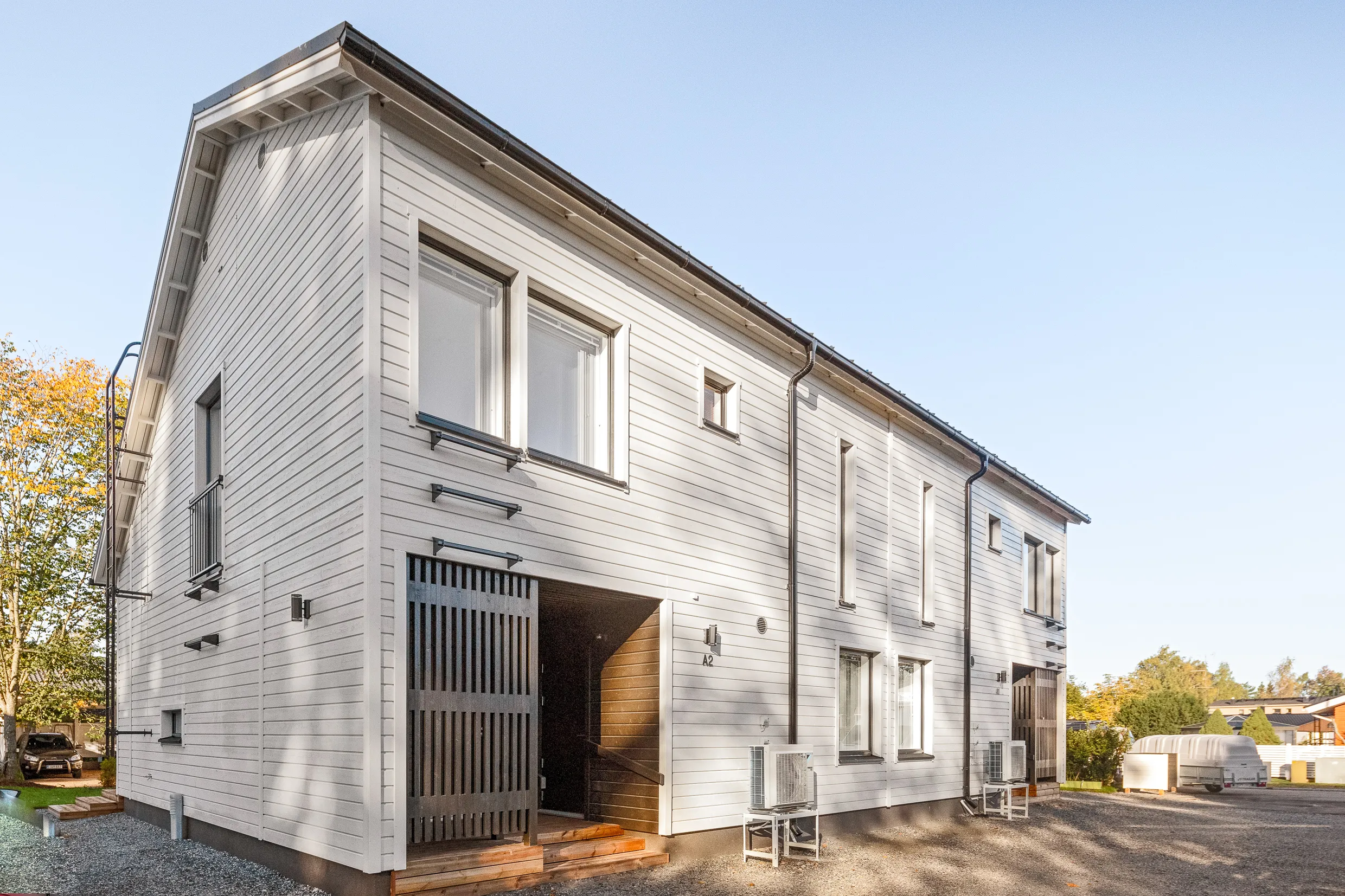 Vaalea, puuverhoiltu rakennus, jossa on tumma liukuovi ja pieniä ikkunoita, valoisassa säässä.