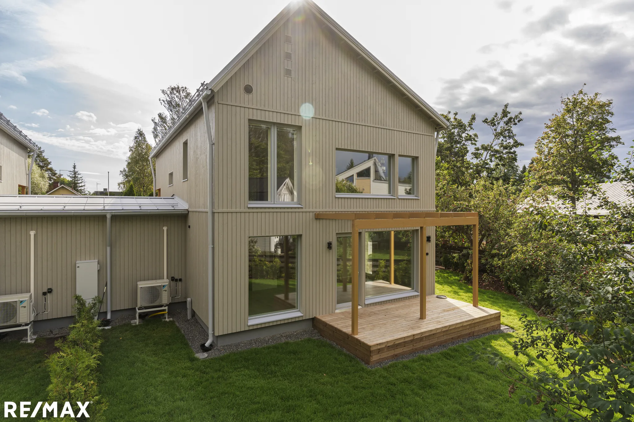 Valokuva modernista, beige-värisestä puutalosta, jossa on suuret ikkunat ja puinen terassi, aurinkoisena päivänä.