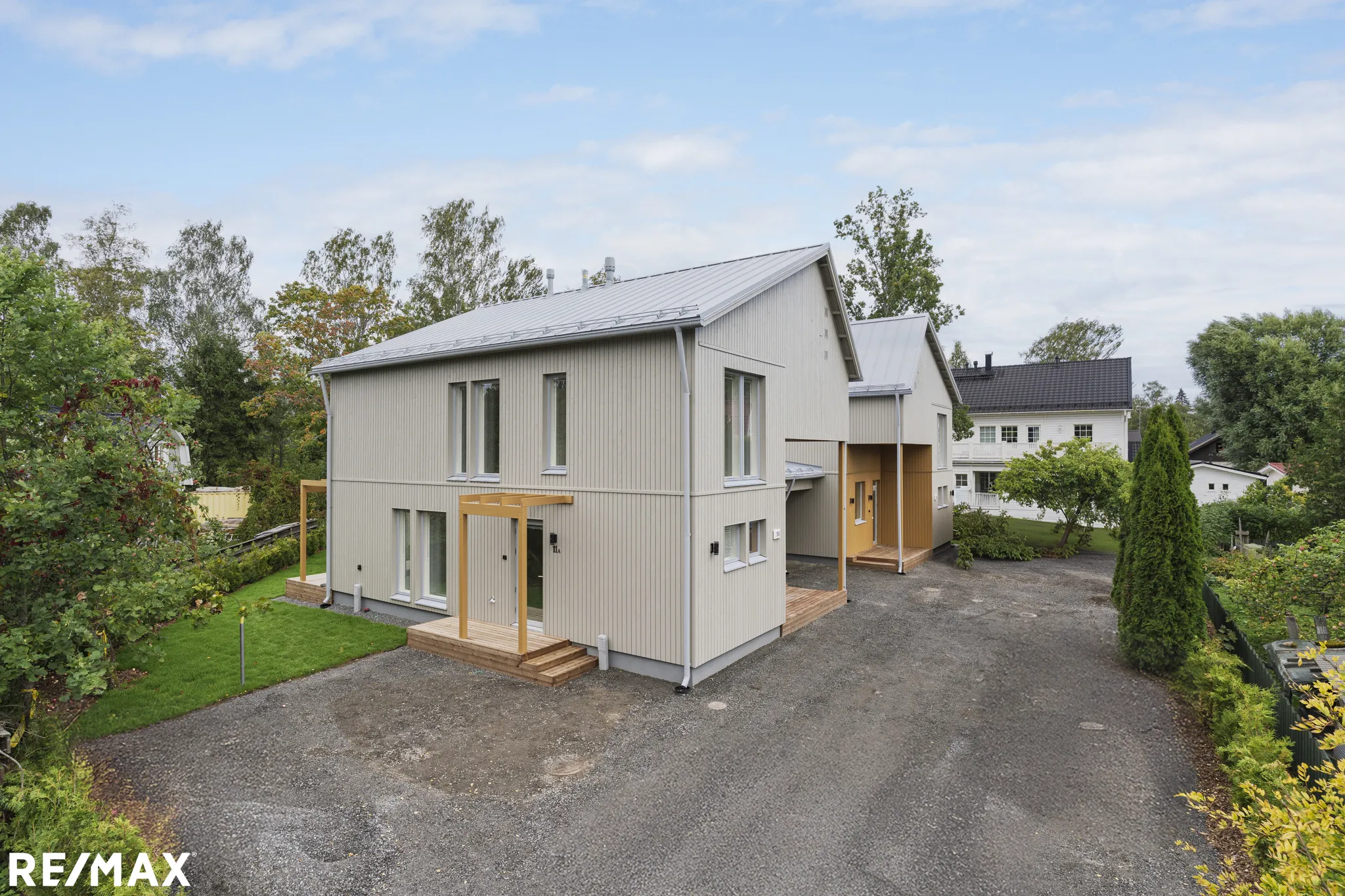 Kaksikerroksinen, vaaleanbeige puutalo, jossa harmaa peltikatto ja puinen terassi, aurinkoisessa säässä.