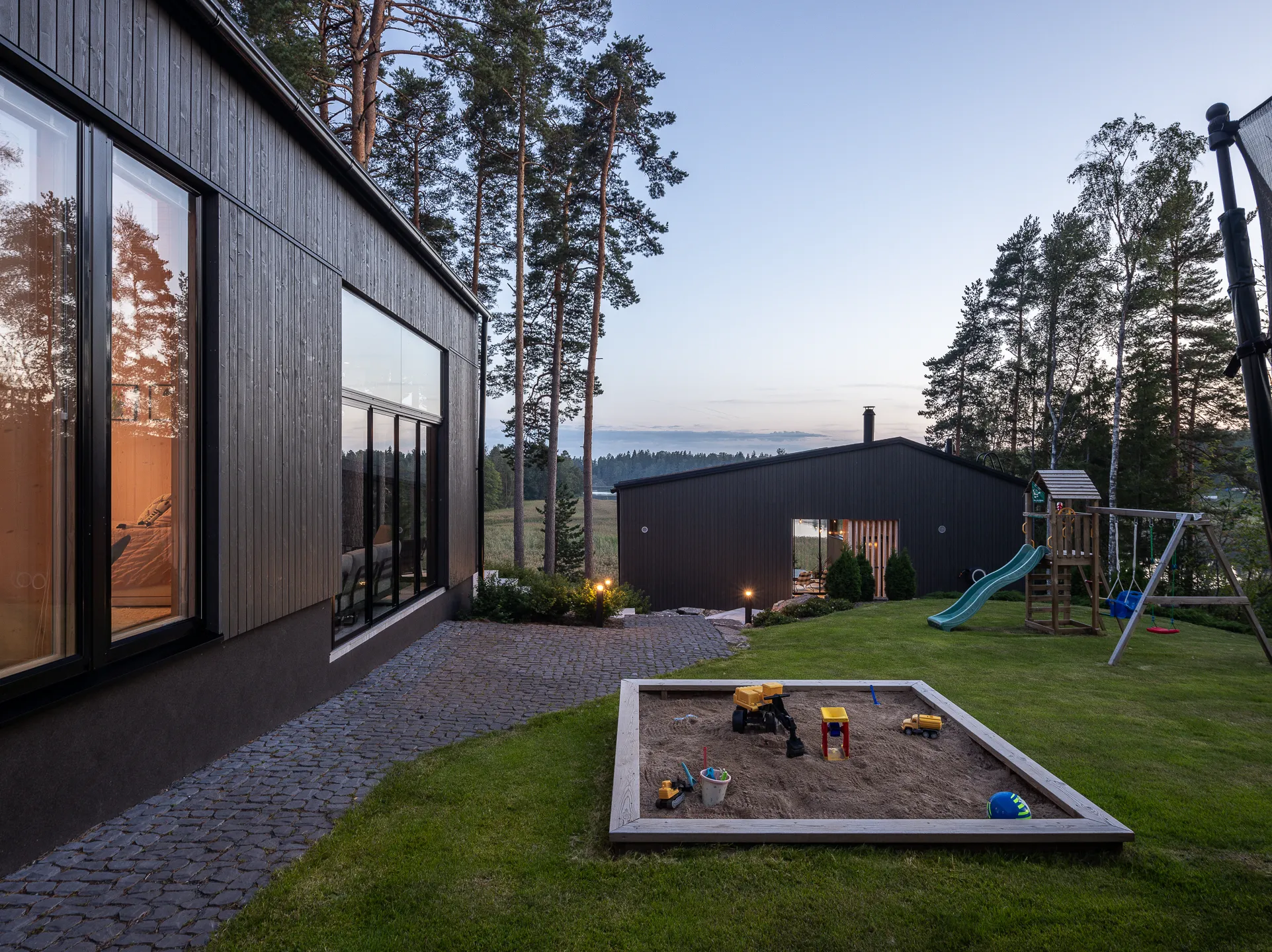 Moderni, tumma puutalo, jossa suuret ikkunat, kivetty polku, hiekkalaatikko leluineen ja leikkikenttä vihreällä nurmikolla iltavalaistuksessa.
