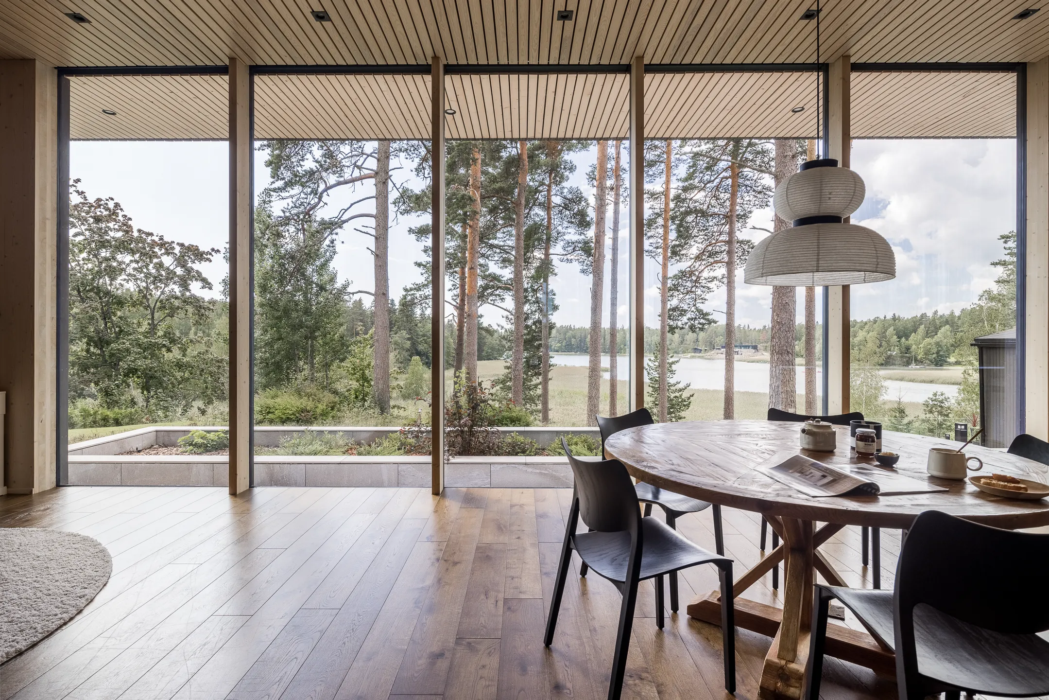 Valoisassa ruokailutilassa on suuri pyöreä puupöytä, mustat tuolit ja suuret ikkunat, joista avautuu näkymä järvelle ja metsään. Katossa on puupanelointi ja riippuvalaisin.