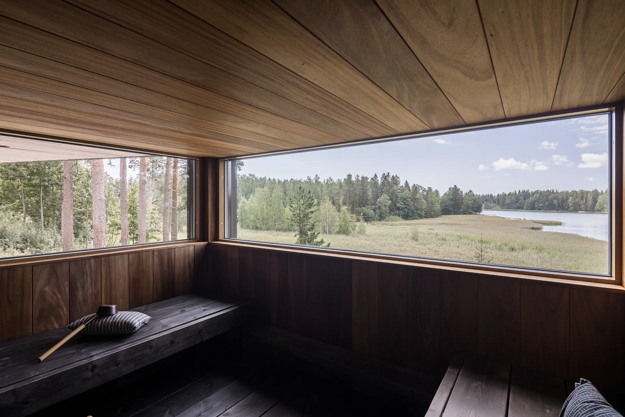 Puupaneloitu sauna, jossa on suuret ikkunat, joista näkyy metsä ja järvi, penkillä tyyny ja kauha.