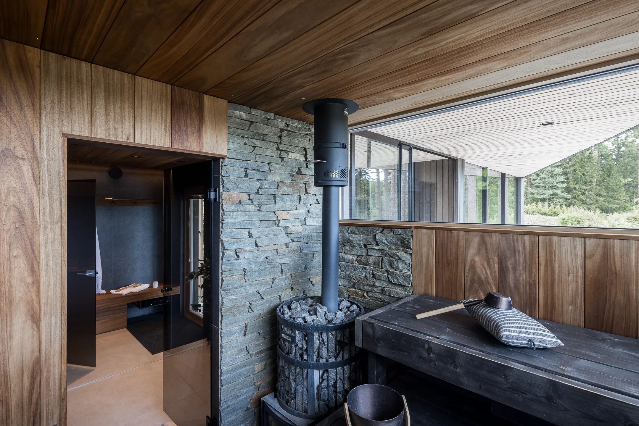 Sisätiloissa on puupaneloitu sauna, jossa on kiviseinä, musta kiuas ja ikkuna, josta näkyy ulos.