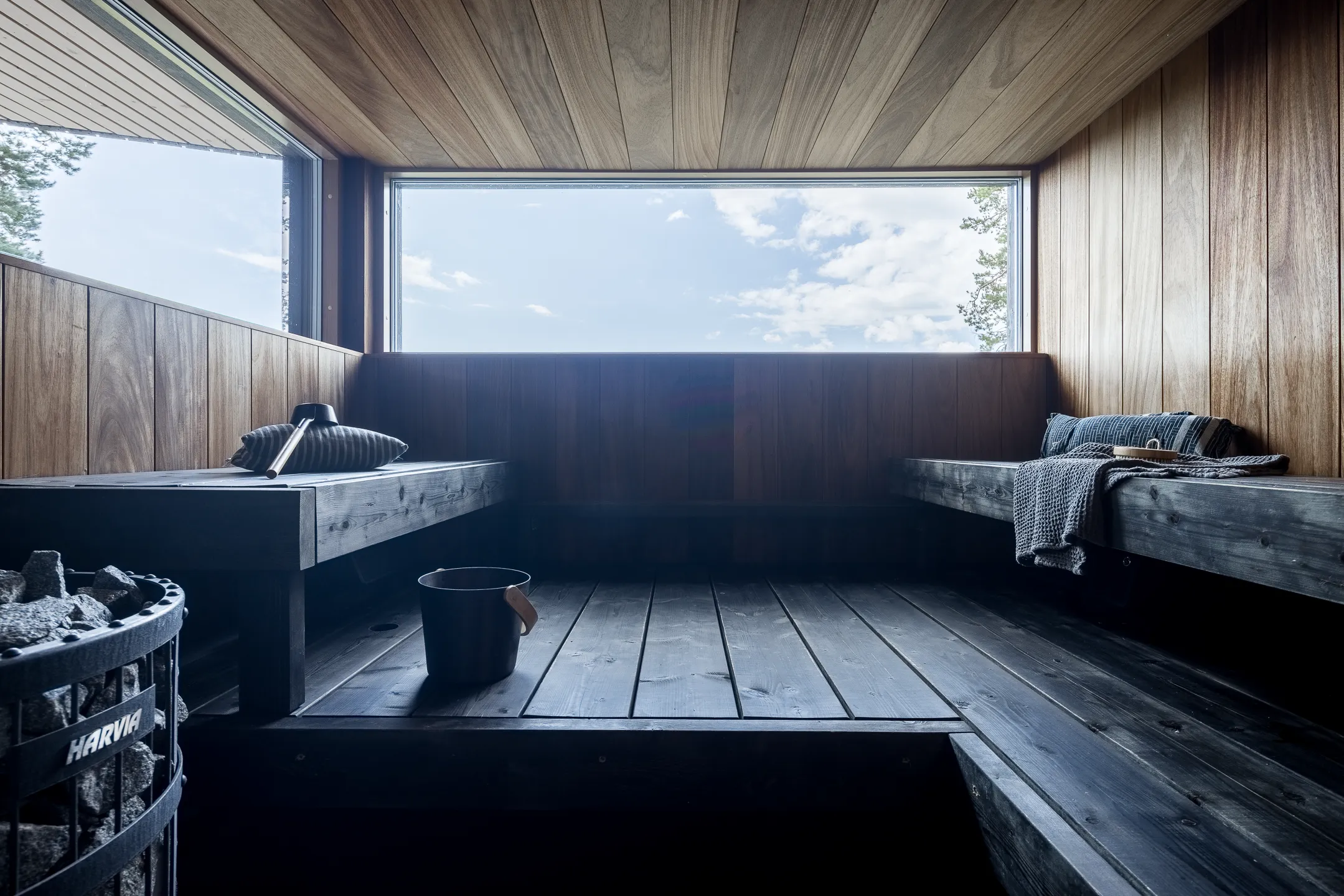 Sauna, jossa on puupanelointi, ikkuna ja kivikiuas. Istuimet ovat tummat ja valaistus on himmeä.