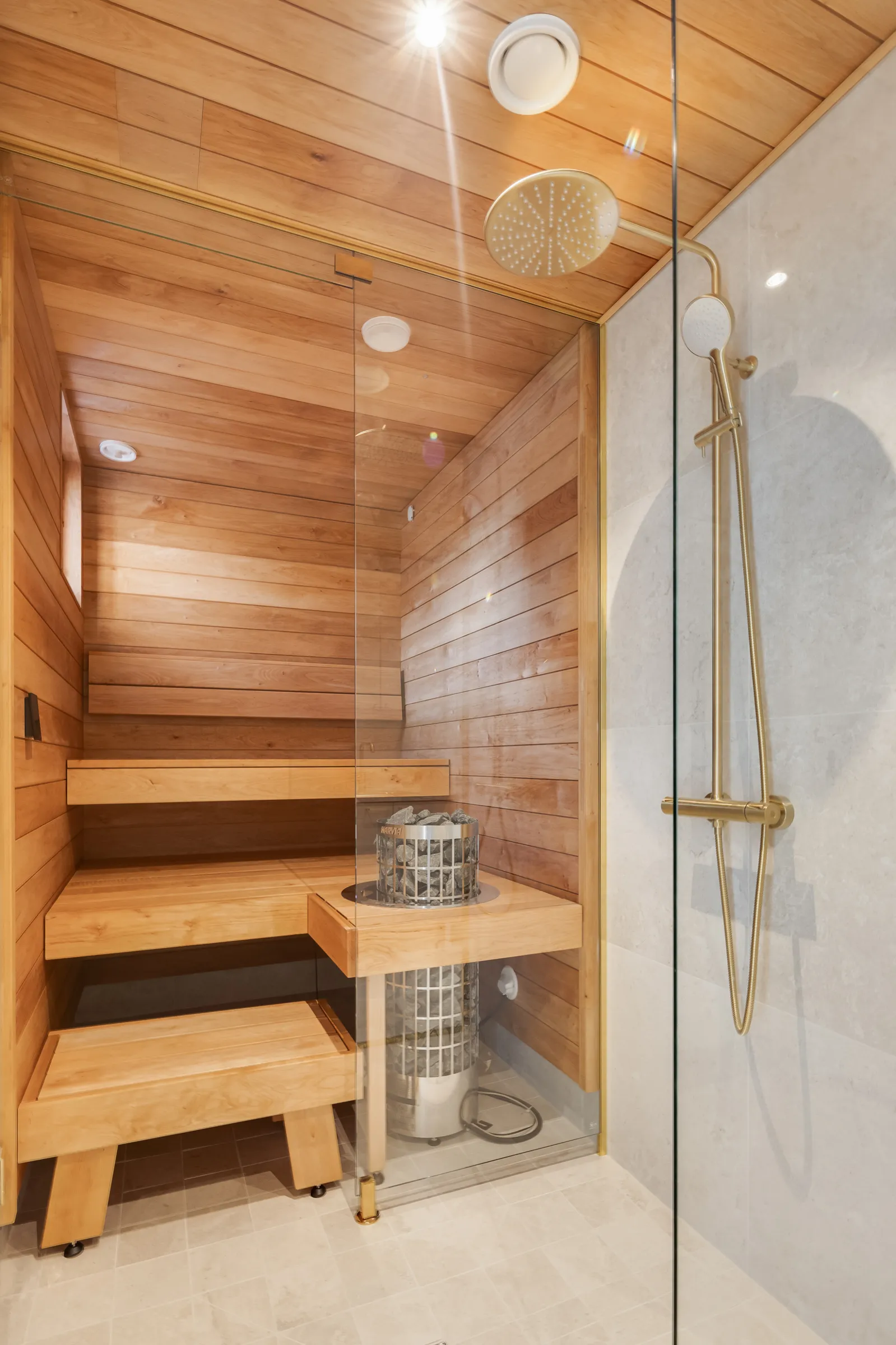 Saunaosasto, jossa on puupanelointi, lasiseinä, puupenkit ja metallinen kiuas kivillä, sekä suihku kullansävyisellä suihkupäällä ja -letkulla.