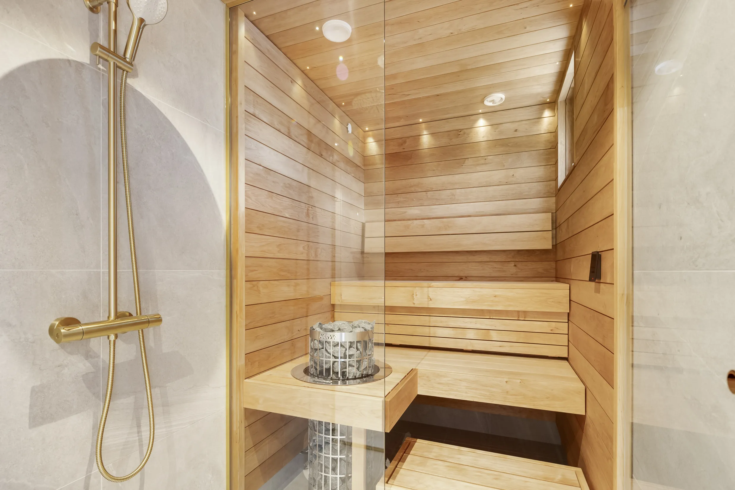 Kuvassa on sauna, jossa on puupanelointi, lasiseinä ja metallinen kiuas kivillä. Saunan vieressä on suihku kultaisella suihkupäällä ja harmaalla seinällä.