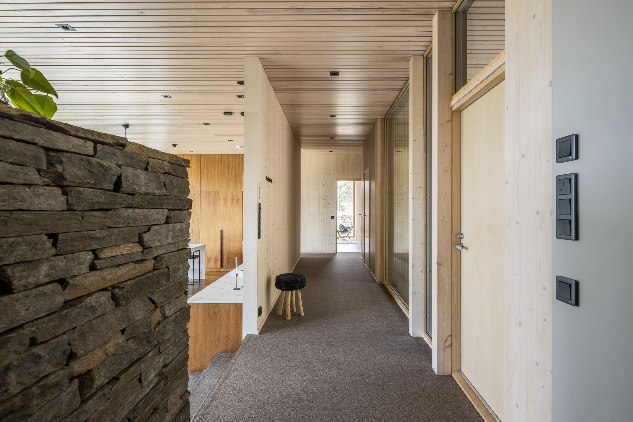 Sisätilakuva kiviseinällä, puukattoisella käytävällä, jossa on harmaa matto, puuseinät ja ulos johtava ovi.