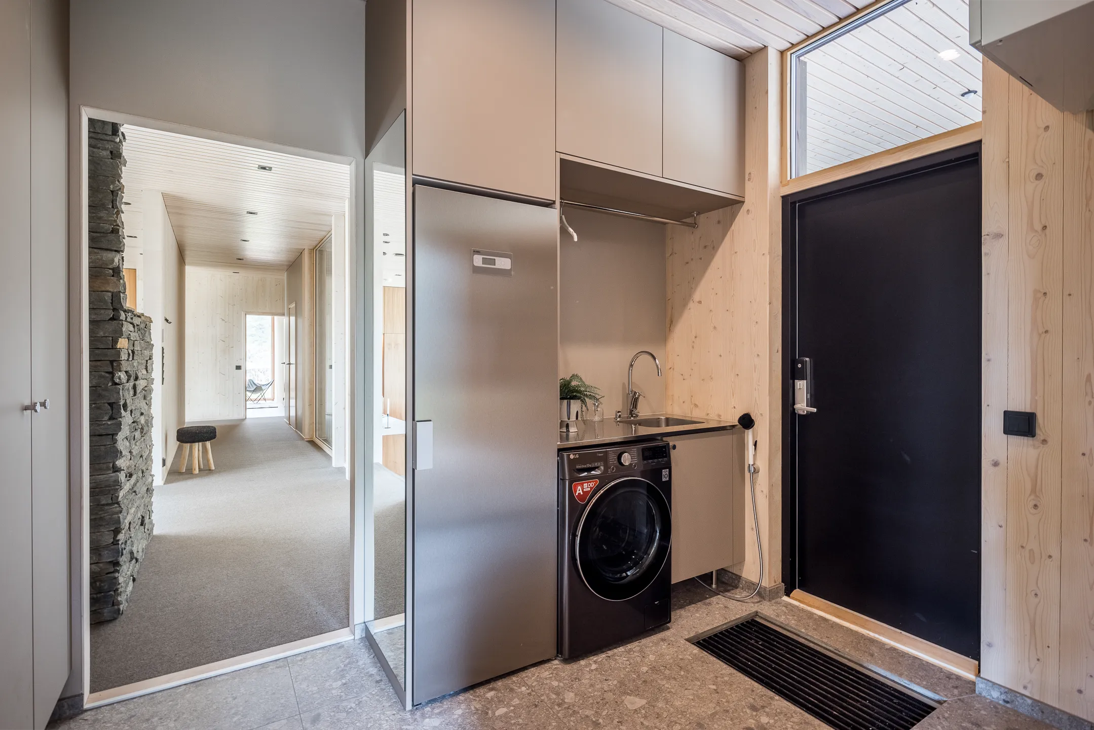Sisätila, jossa on pesukone, pesuallas, kaapit ja ovi, valaistus on kirkas ja värit ovat neutraaleja.
