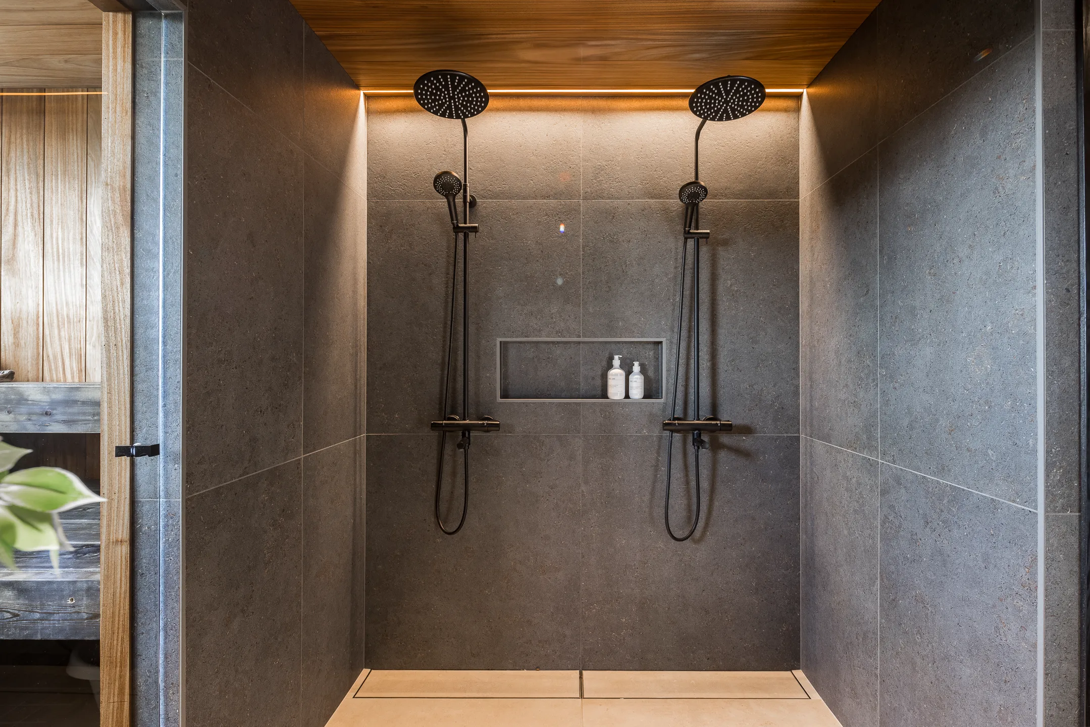Moderni kylpyhuone, jossa kaksi mustaa suihkua, harmaat laatat ja puinen katto lämpimällä valaistuksella.