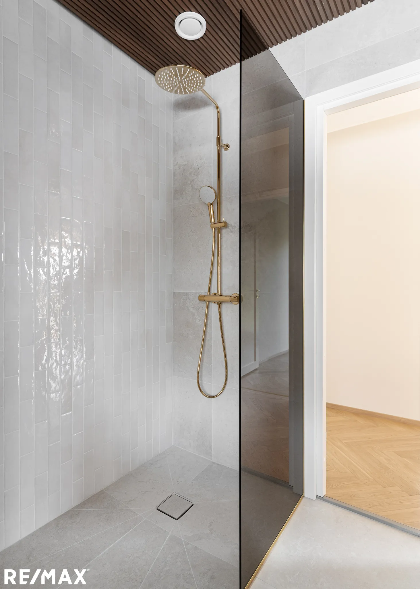 Moderni suihku kultaisella suihkupäällä, valkoisilla seinälaatoilla ja harmaalla lattialla, jossa on RE/MAX-logo alhaalla.