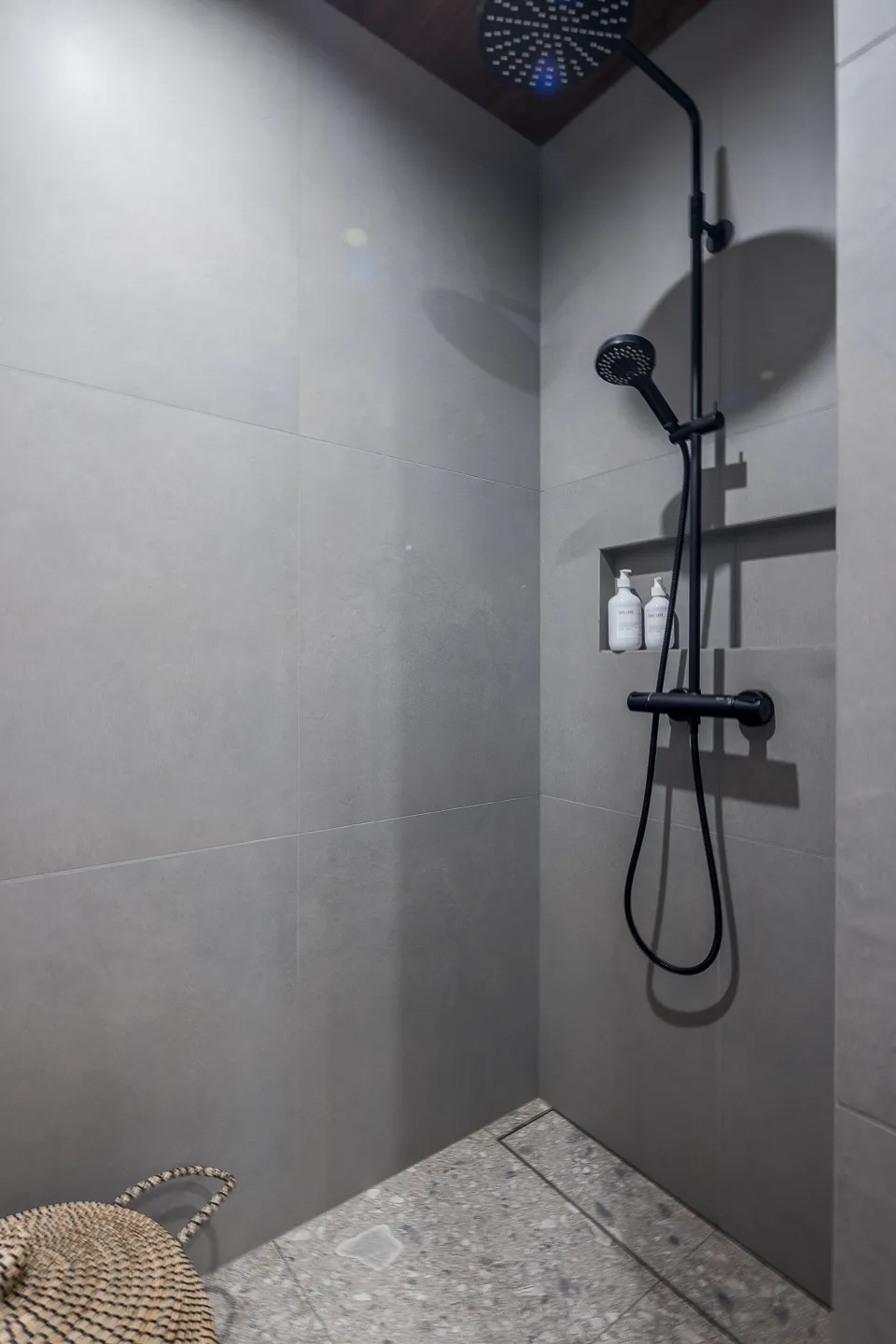 Moderni suihkunurkkaus harmailla laatoilla, mustalla suihkusetillä ja sisäänrakennetulla hyllyllä, jossa on kaksi valkoista pulloa. Lattialla on harmaa mosaiikkikuvio ja korikori.