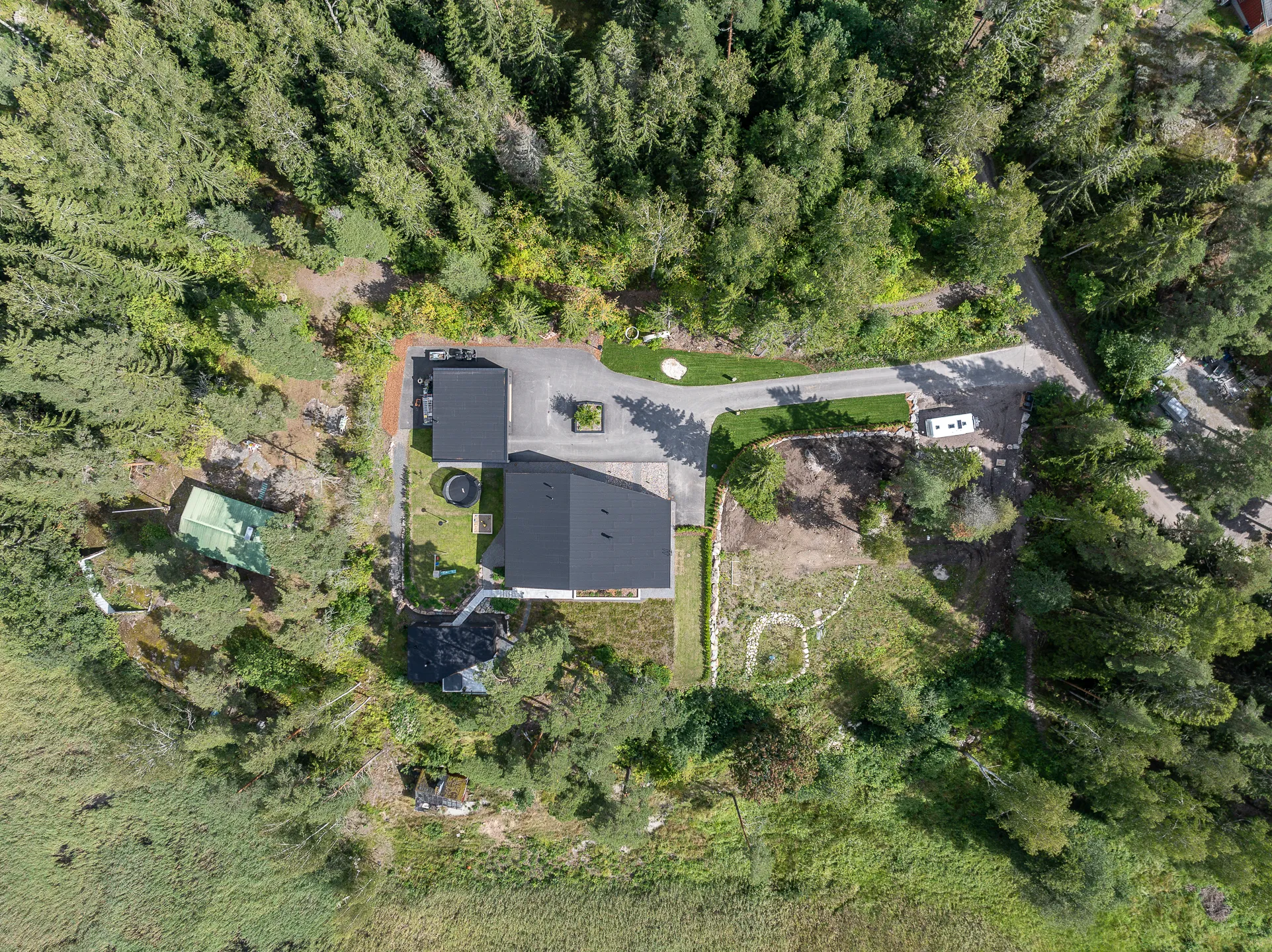 Ylhäältä otettu ilmakuva, jossa näkyy talo, metsä ja niitty. Talolla on tumma katto ja se sijaitsee metsän ja niityn välissä. Metsä on tiheä ja vihreä, ja niitty on avoin ja ruohoinen. Valo on kirkas ja aurinkoinen.