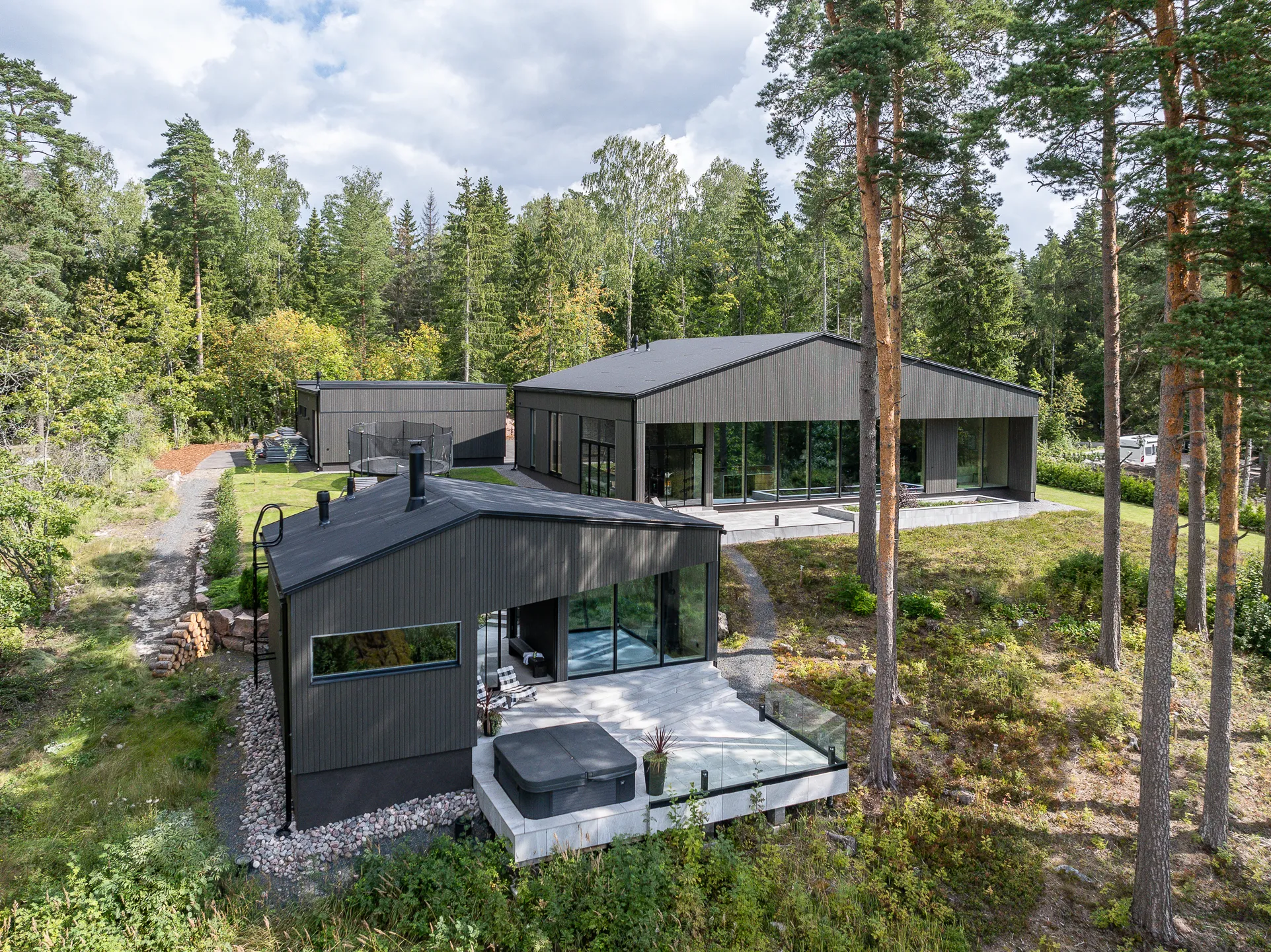 Moderni, tummanharmaa talo metsäisessä ympäristössä, jossa on suuret ikkunat, terassi ja poreallas.