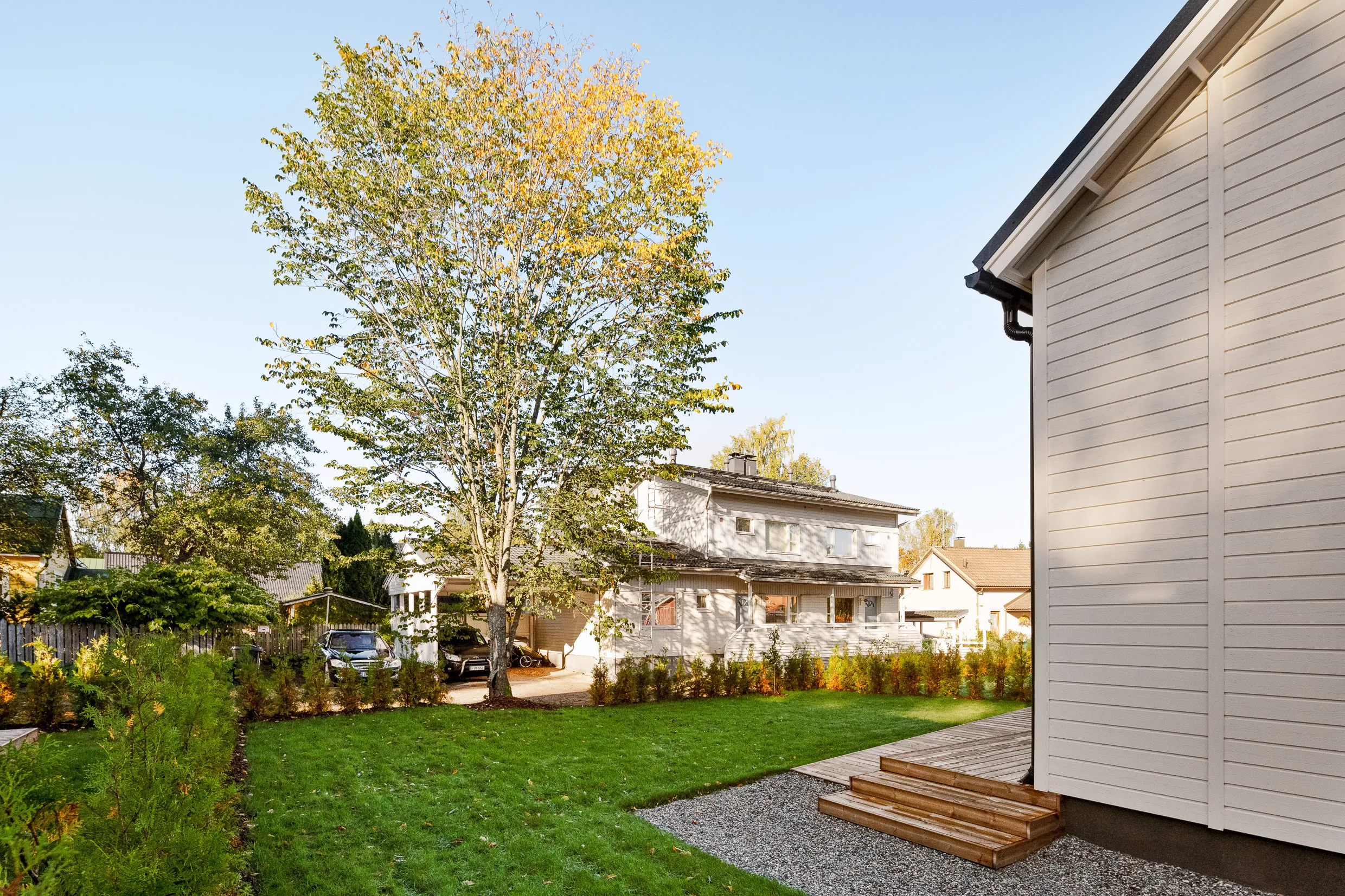 Ulkonäkymä valkoisesta talosta, jossa on vihreä nurmikko, puita ja harmaa kivipolku aurinkoisena päivänä.
