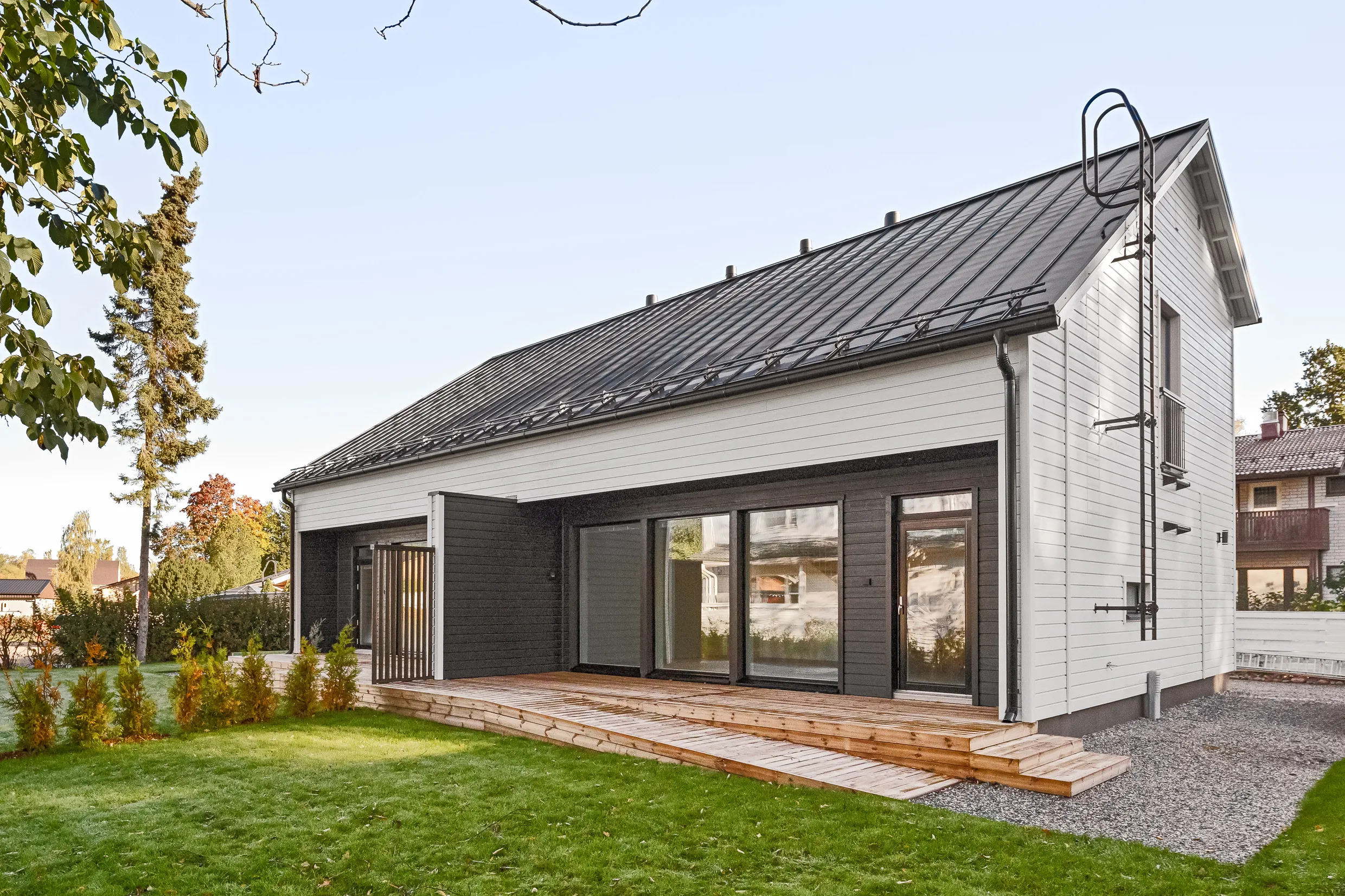 Moderni, kaksikerroksinen talo, jossa on tumma katto, valkoiset seinät ja puinen terassi. Etualalla on vihreä nurmikko ja pieniä puita.