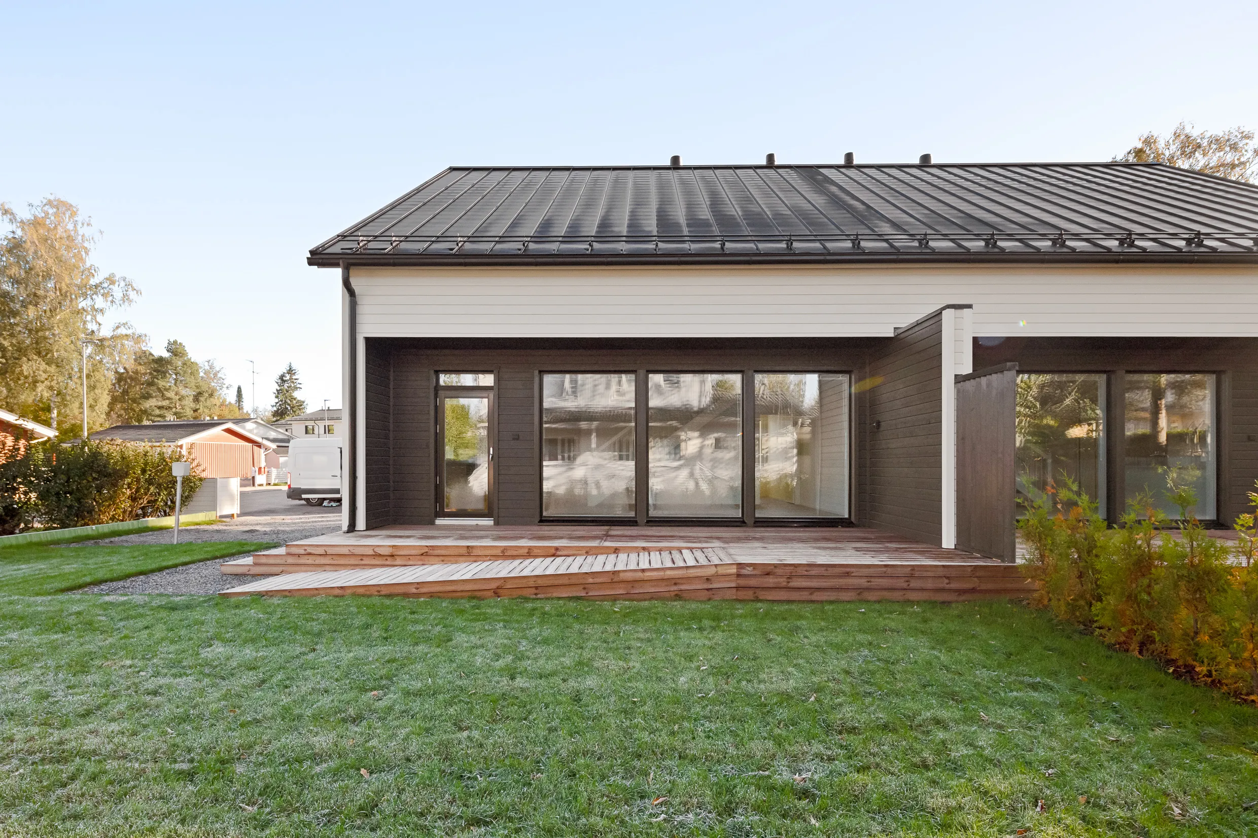 Moderni talo, jossa on suuri puinen terassi, tumma katto ja vihreä nurmikko aamukasteella.