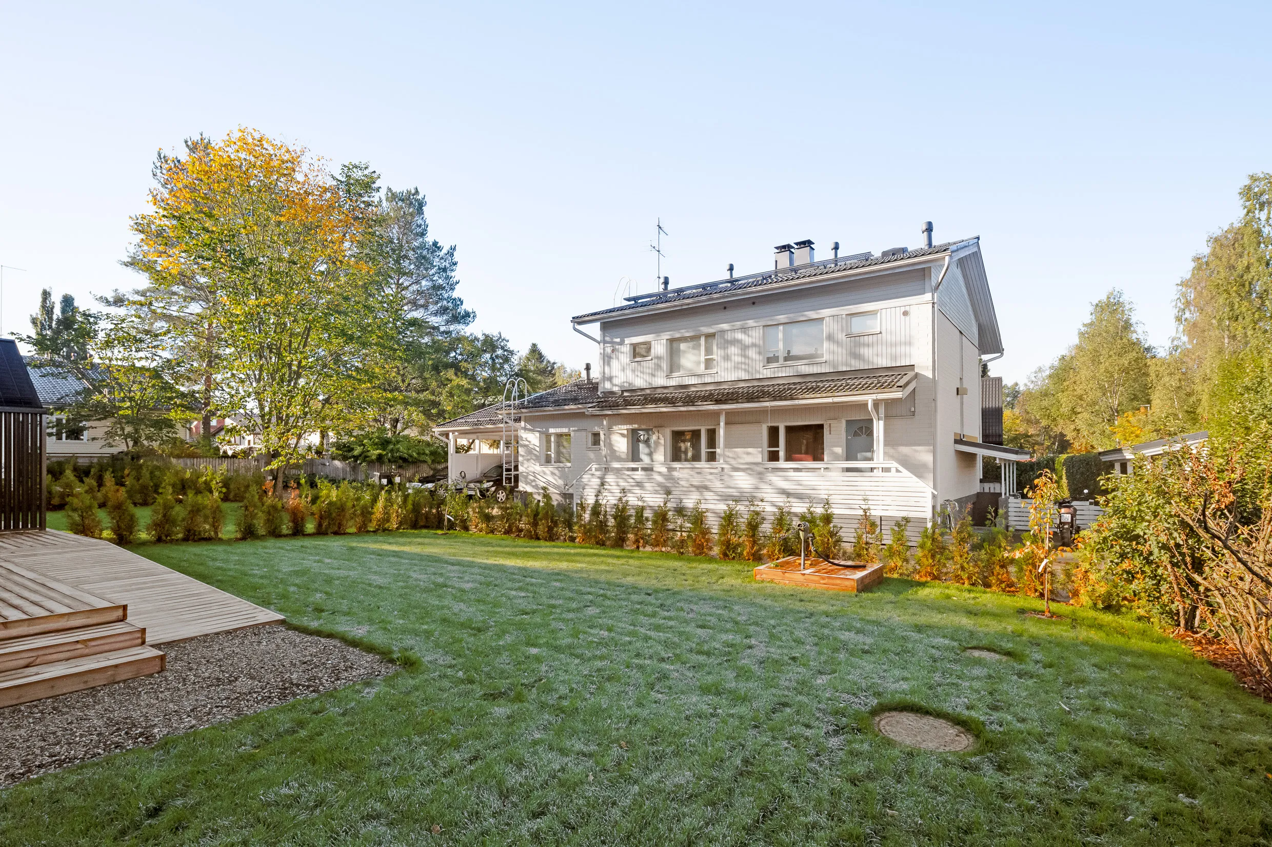 Kaksikerroksinen vaalea puutalo, jossa on vihreä nurmikko, puinen terassi ja syksyinen puu auringonpaisteessa.