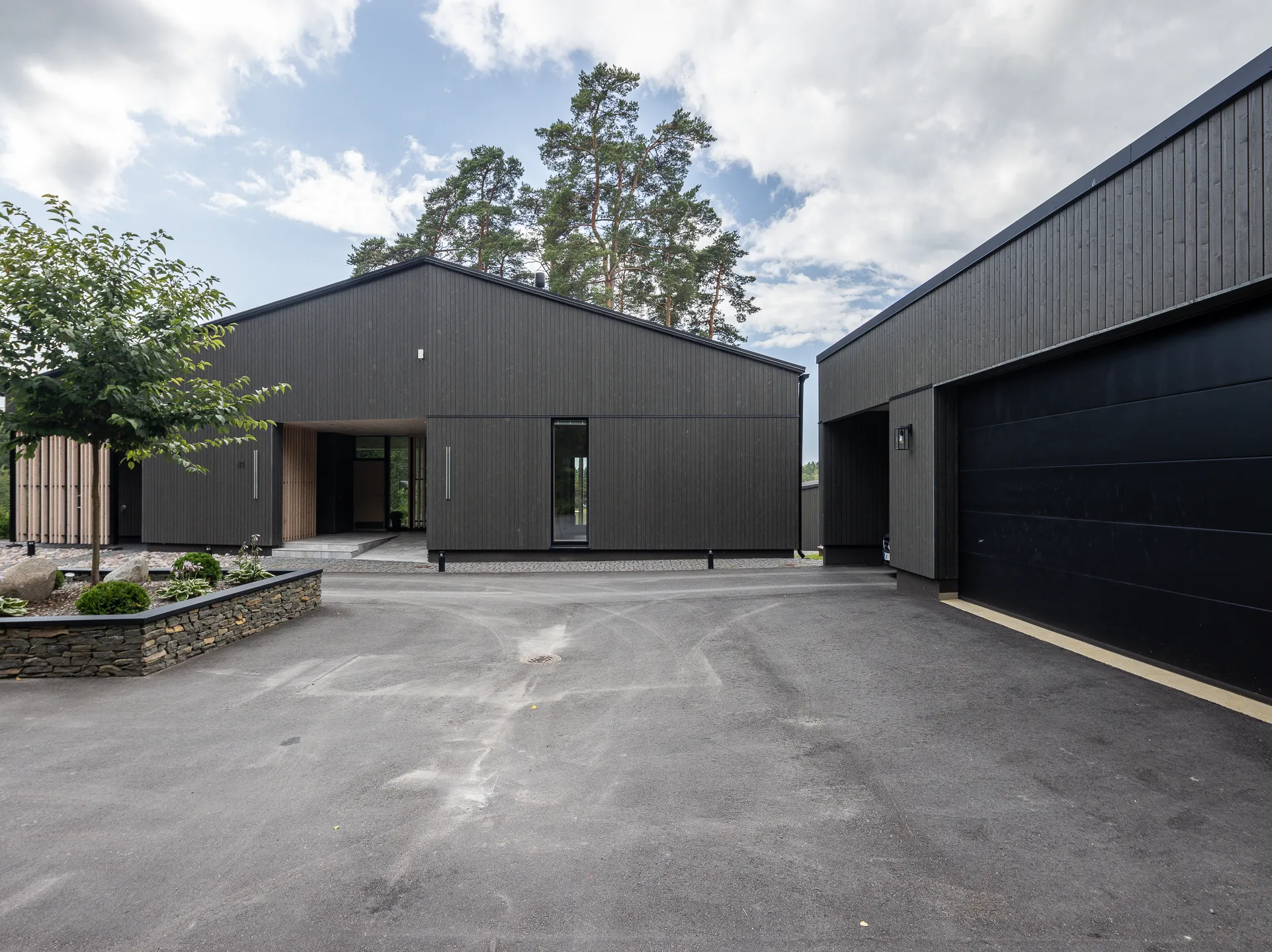 Moderni, tumma puutalo, jossa on musta autotalli ja asfaltoitu piha, valoisassa säässä.