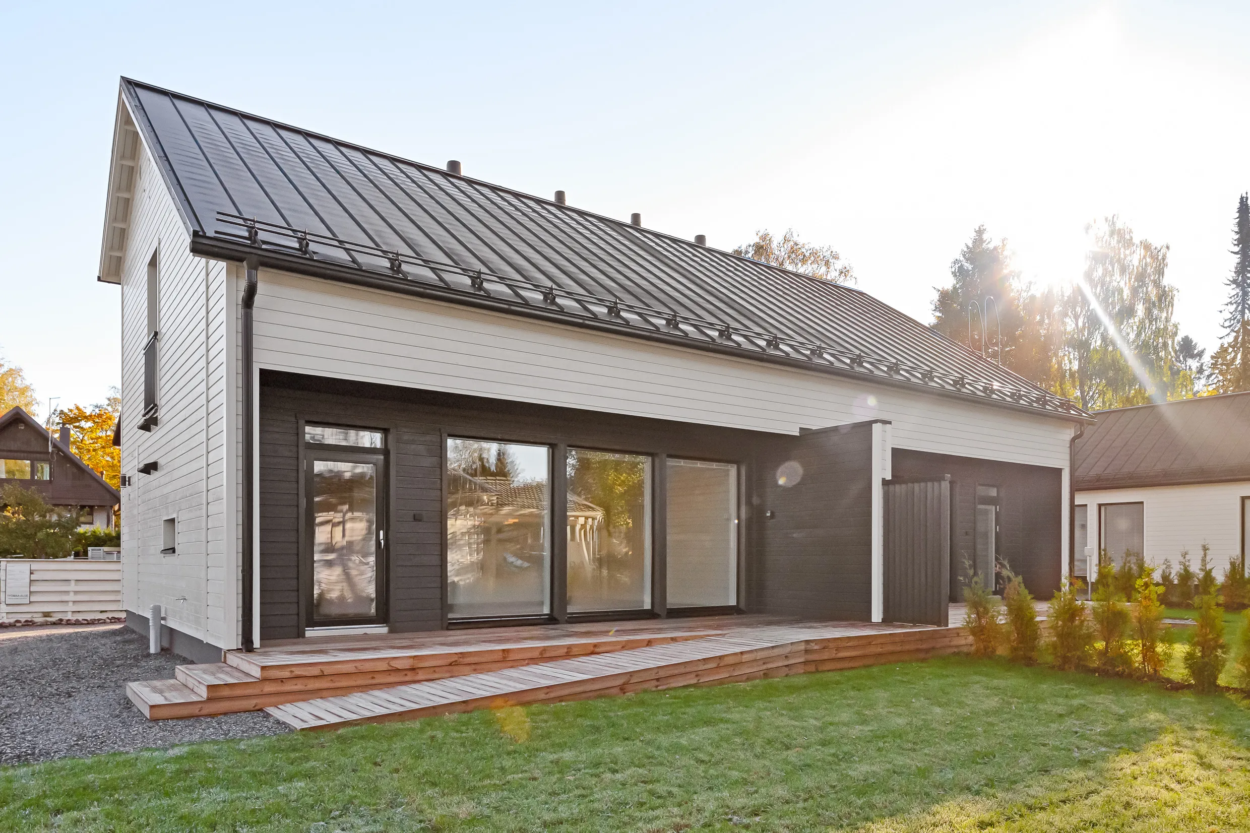 Moderni, yksikerroksinen omakotitalo, jossa on tumma metallikatto, vaaleat seinät ja tummat puuosat, sekä puinen terassi ja vihreä nurmikko aurinkoisena päivänä.